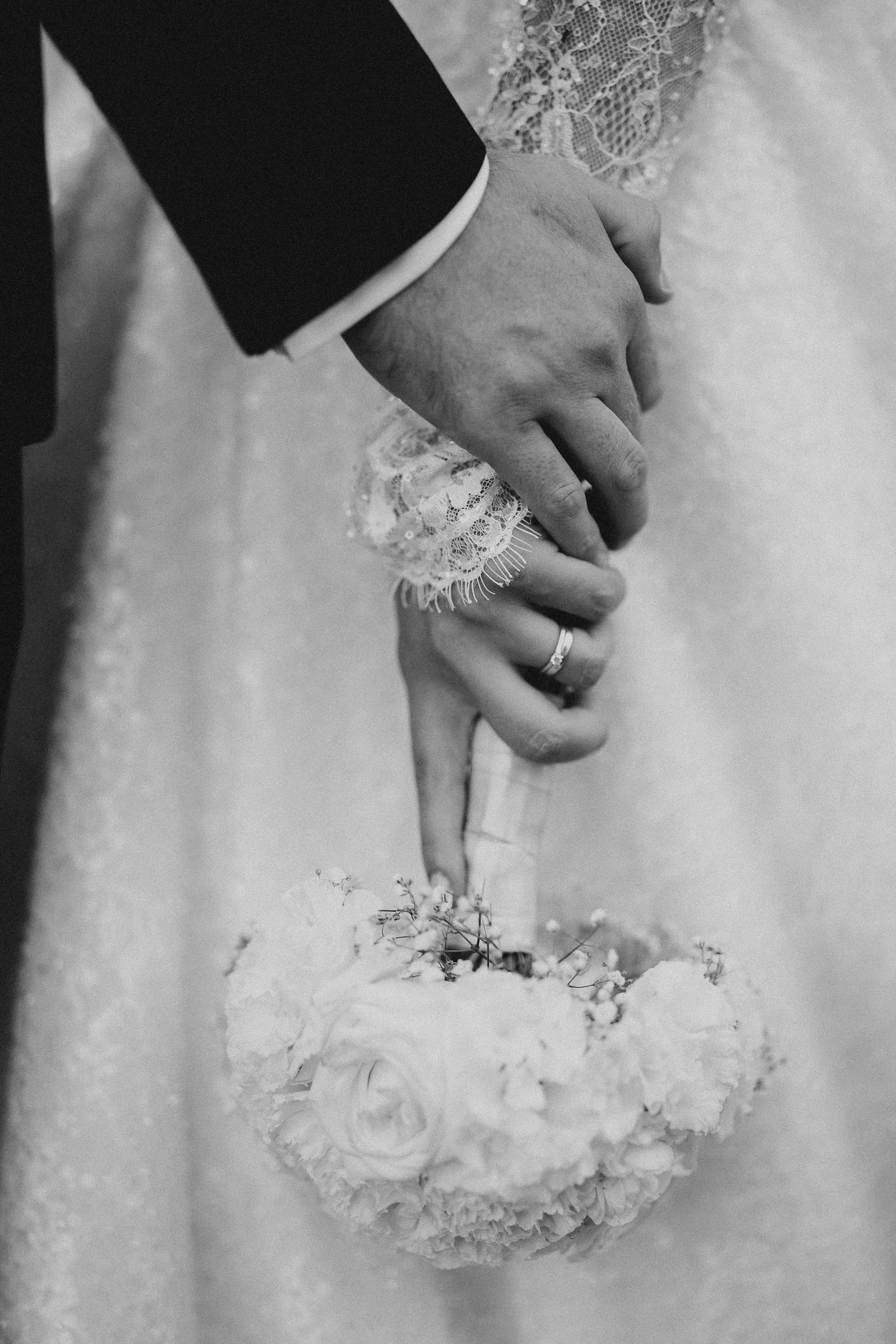 Close-up black and white photo of a person's hand gently touching a lace fabric with their thumb, while the other hand is in the background, with a wedding ring visible on the ring finger.
