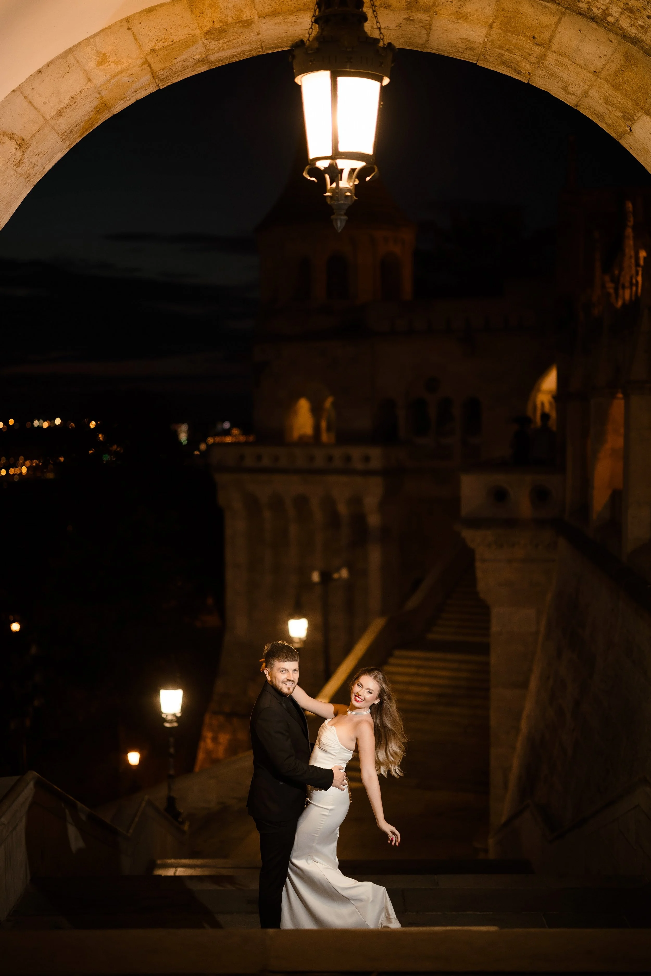 A couple in formal wedding attire dancing on an outdoor staircase at night under a streetlamp, with historic buildings and city lights in the background.