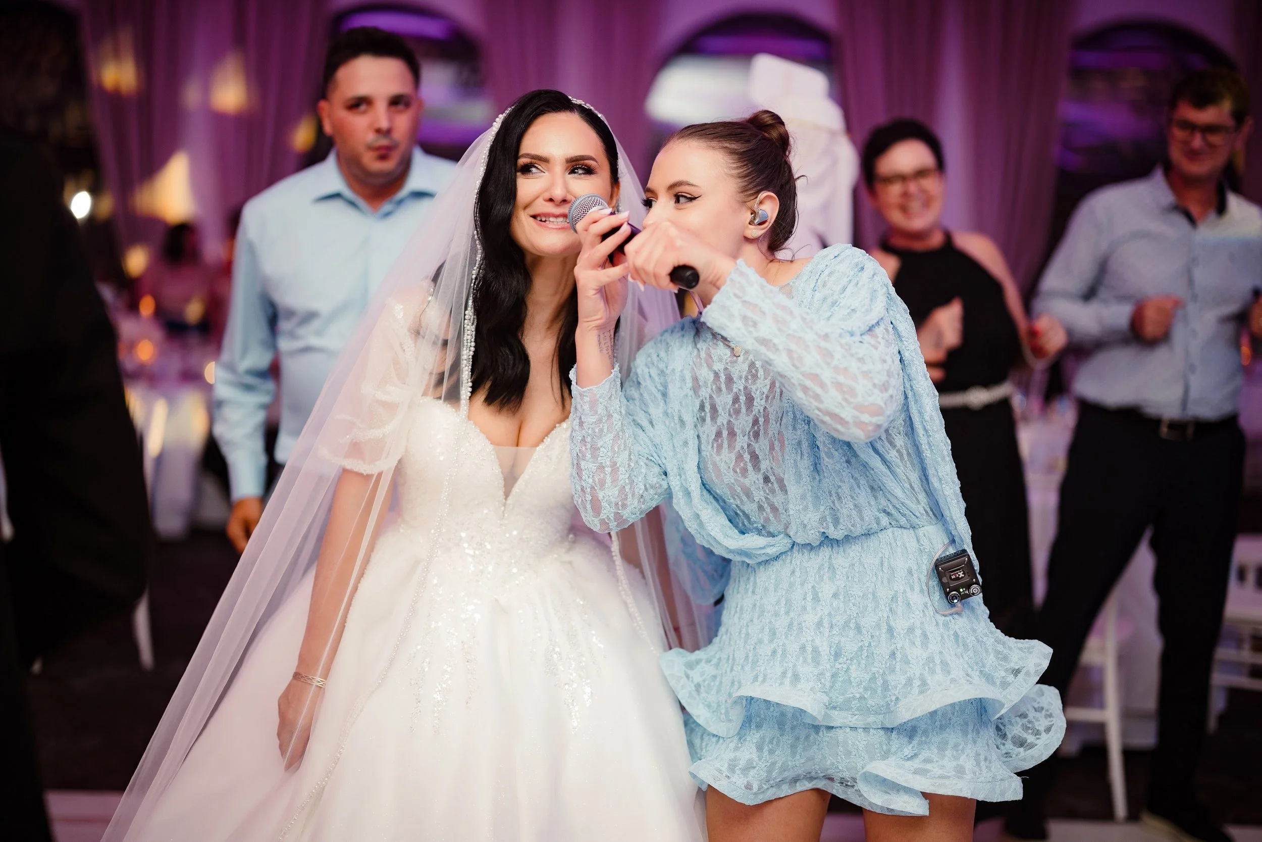 A bride in a white wedding dress and veil is singing into a microphone held by a woman in a blue lace dress, at a wedding reception with guests in the background.
