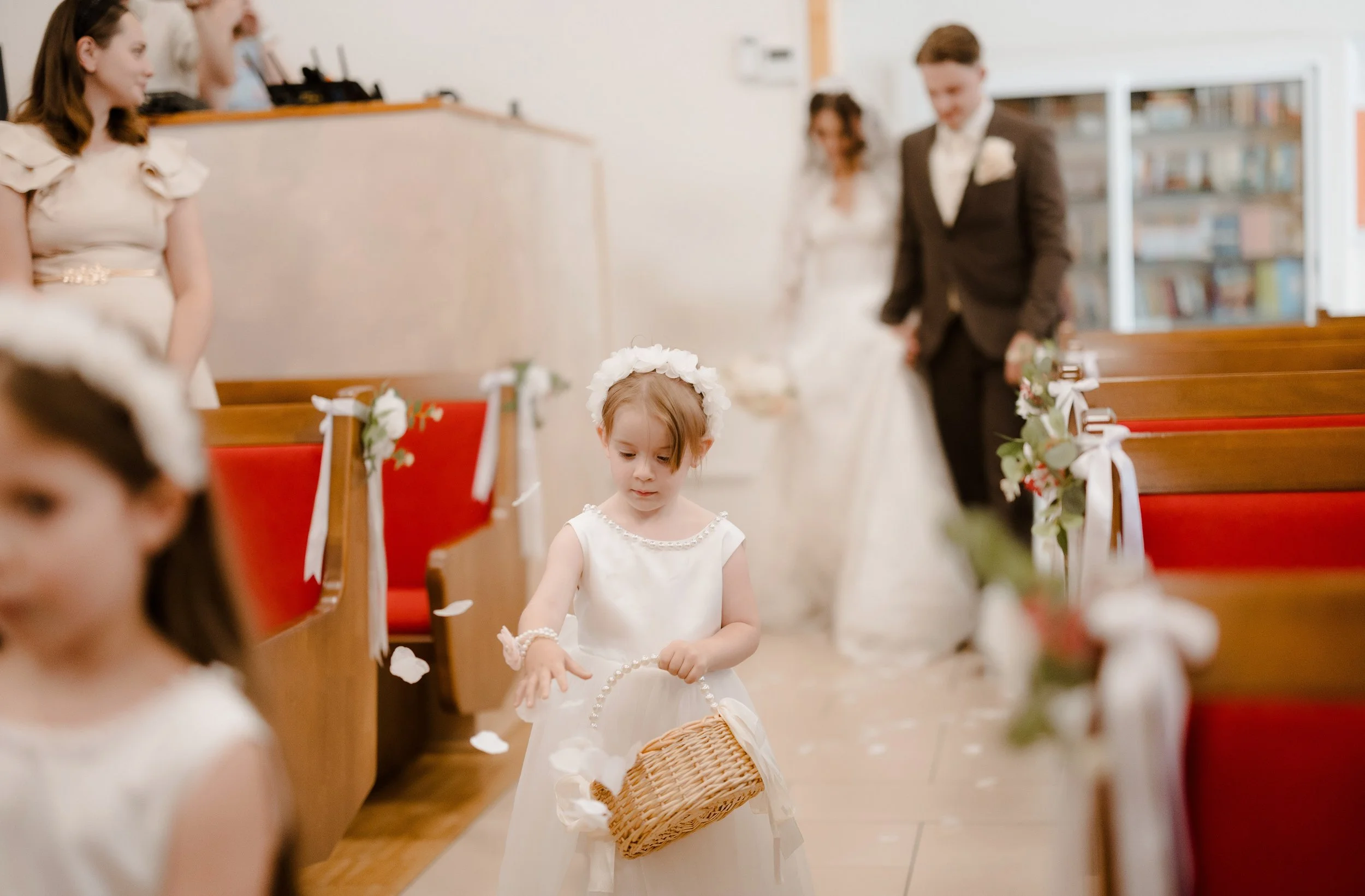 A young girl dressed in a white dress and floral headband collecting flower petals or confetti at a wedding ceremony.