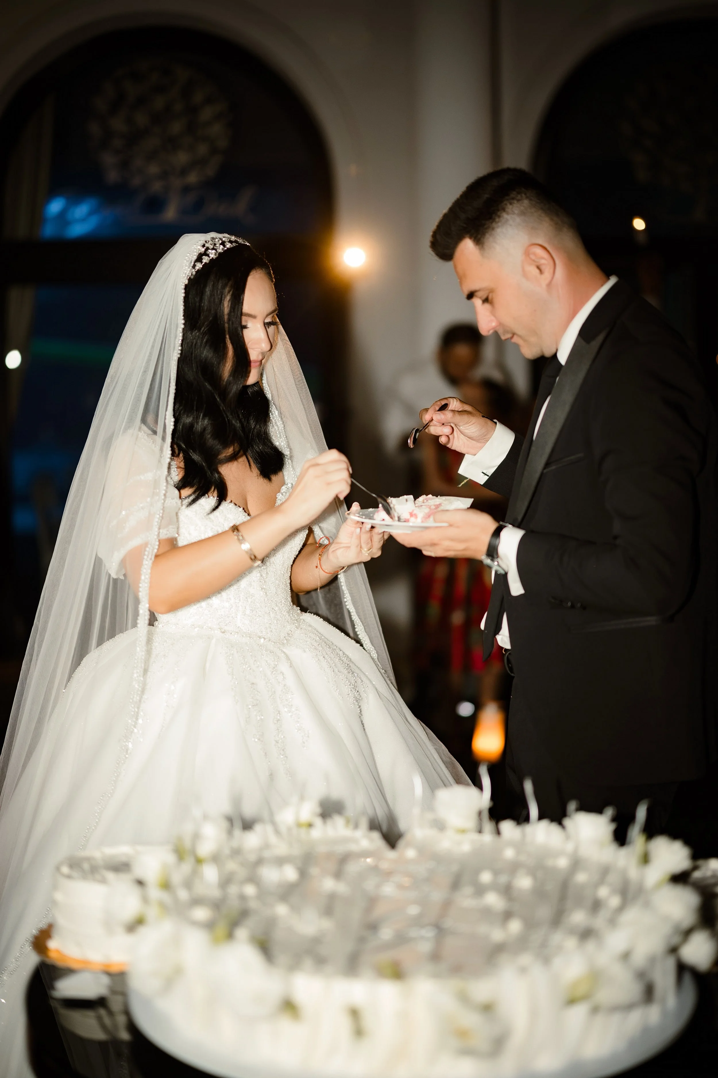 A bride and groom in formal attire sharing a cake at their wedding reception, with a wedding cake in the foreground.