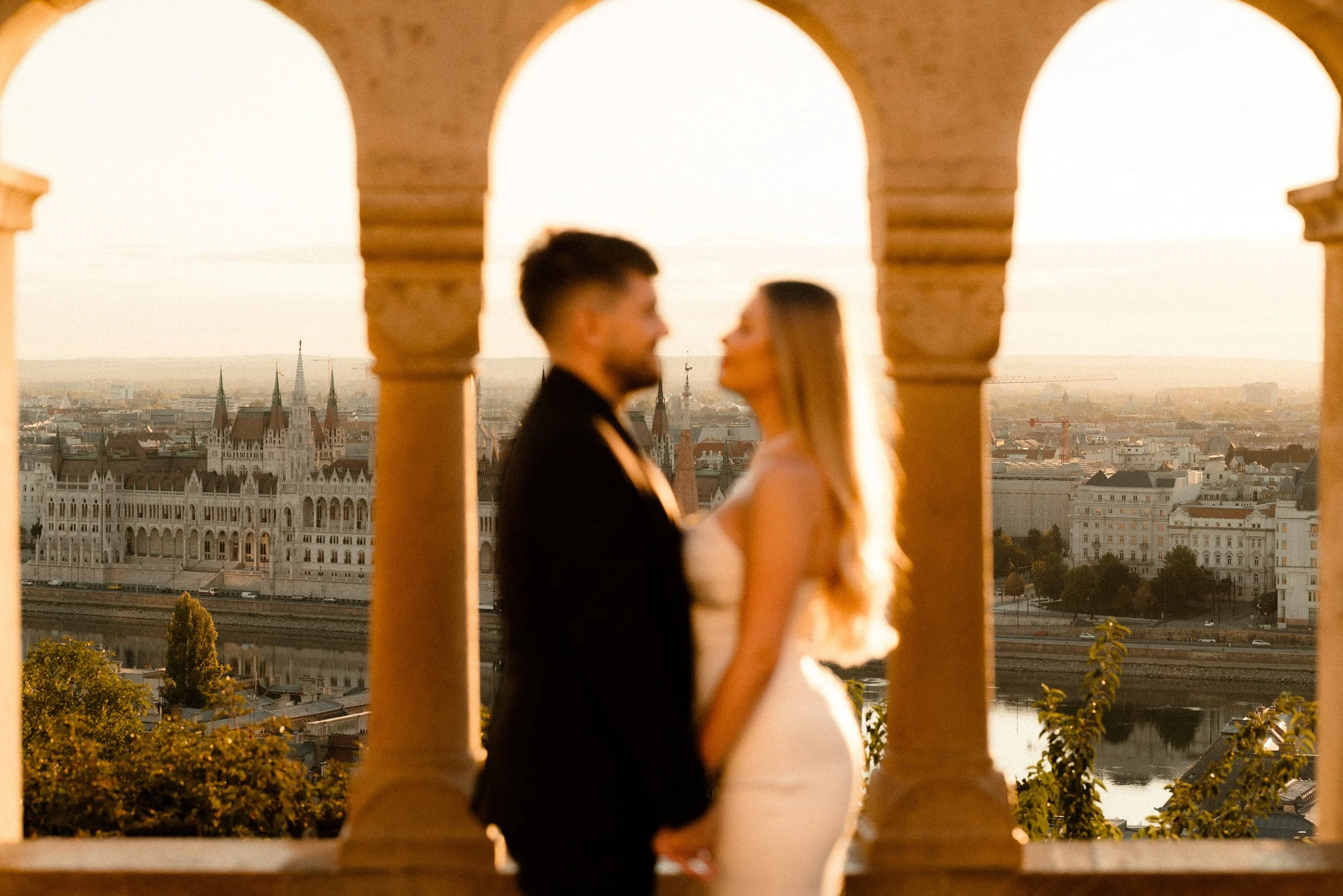 A couple dressed in wedding attire, holding hands and facing each other, captured during sunset on a rooftop with a city skyline in the background.