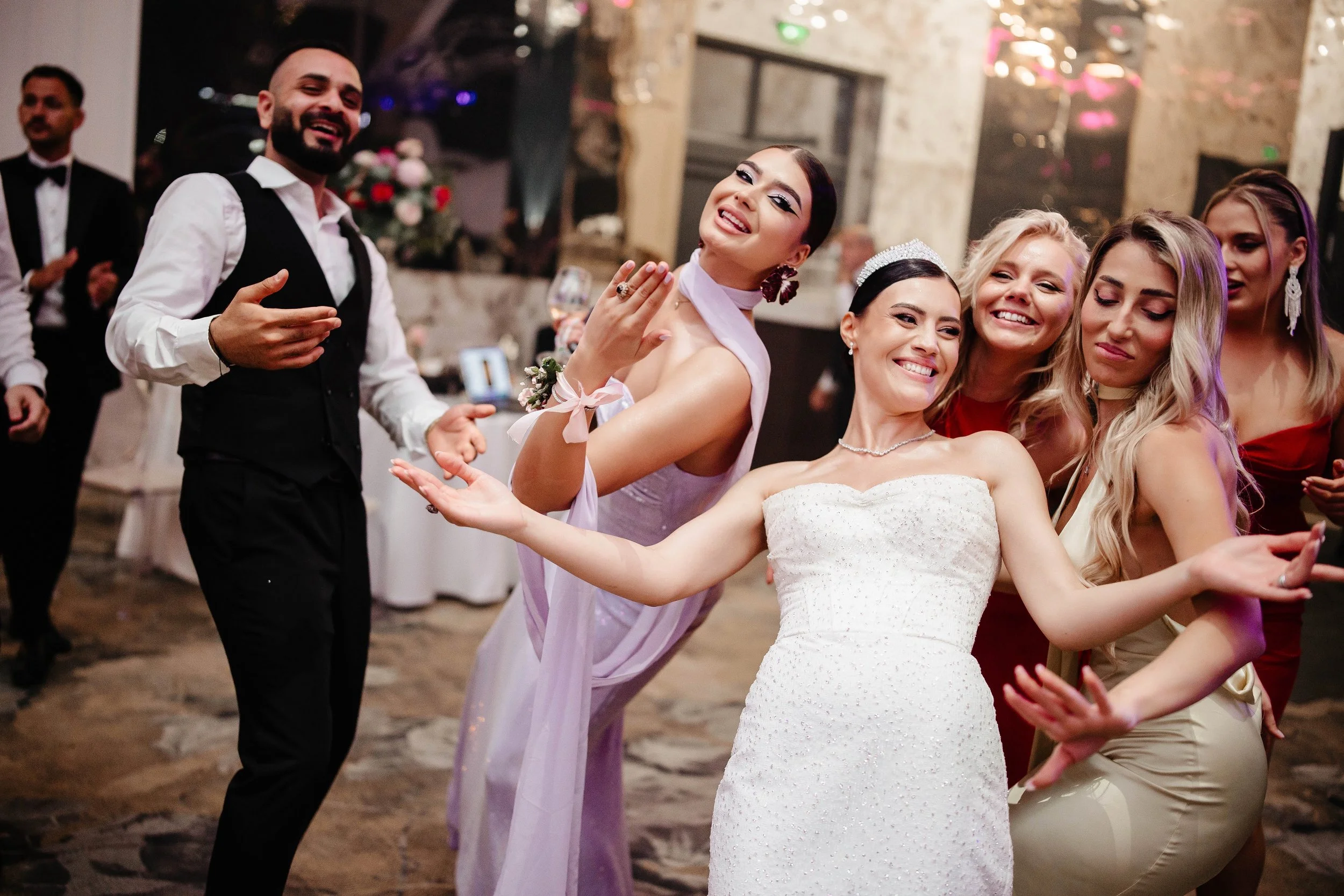 People dancing and celebrating at a wedding reception, with the bride in a white dress and several women in colorful dresses, and a man in a black vest and white shirt.