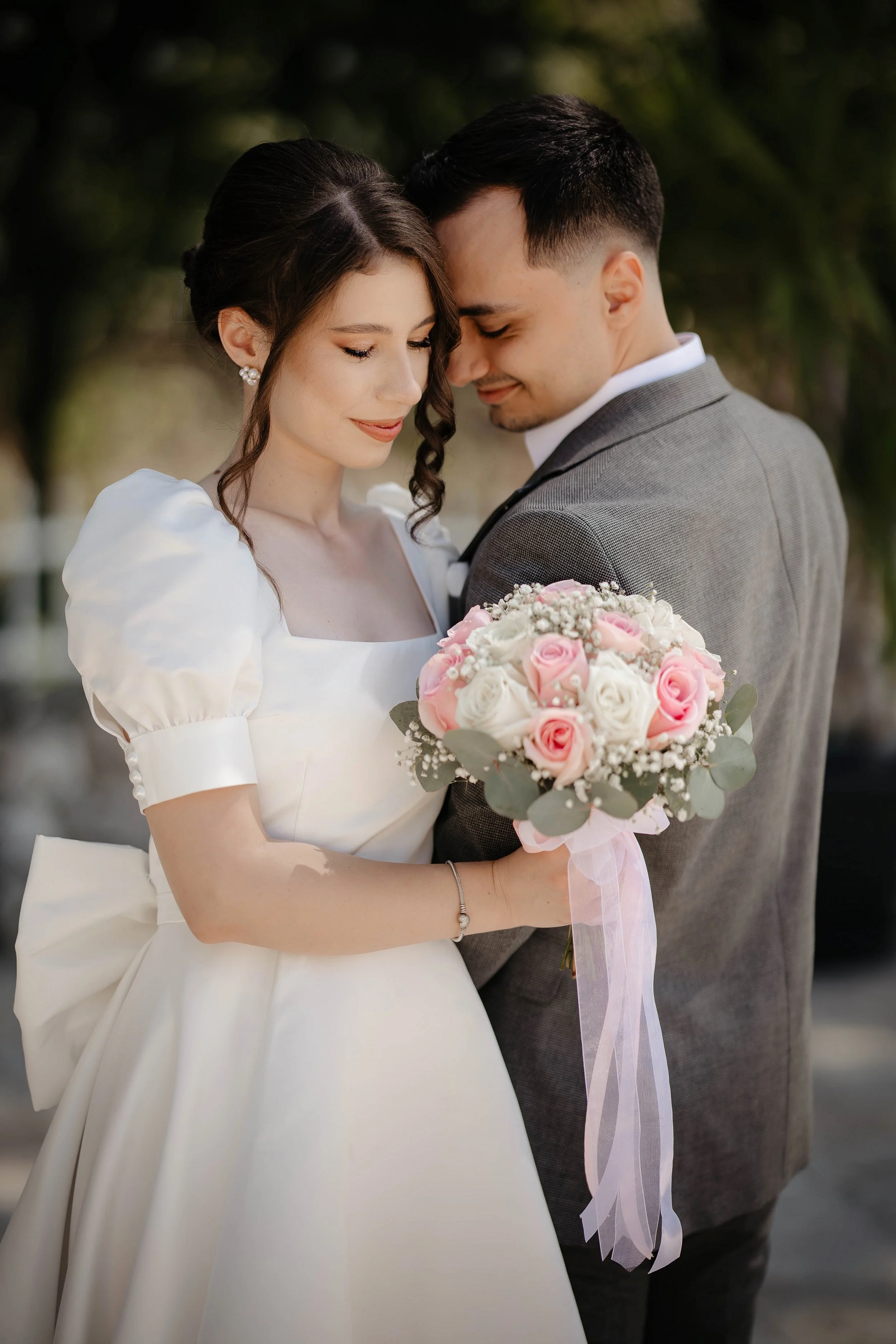 A bride and groom stand close together, smiling with their foreheads touching. The bride wears a white dress with puffed sleeves and has dark, curly hair styled elegantly. The groom wears a gray suit with a white shirt, and they are holding a bouquet