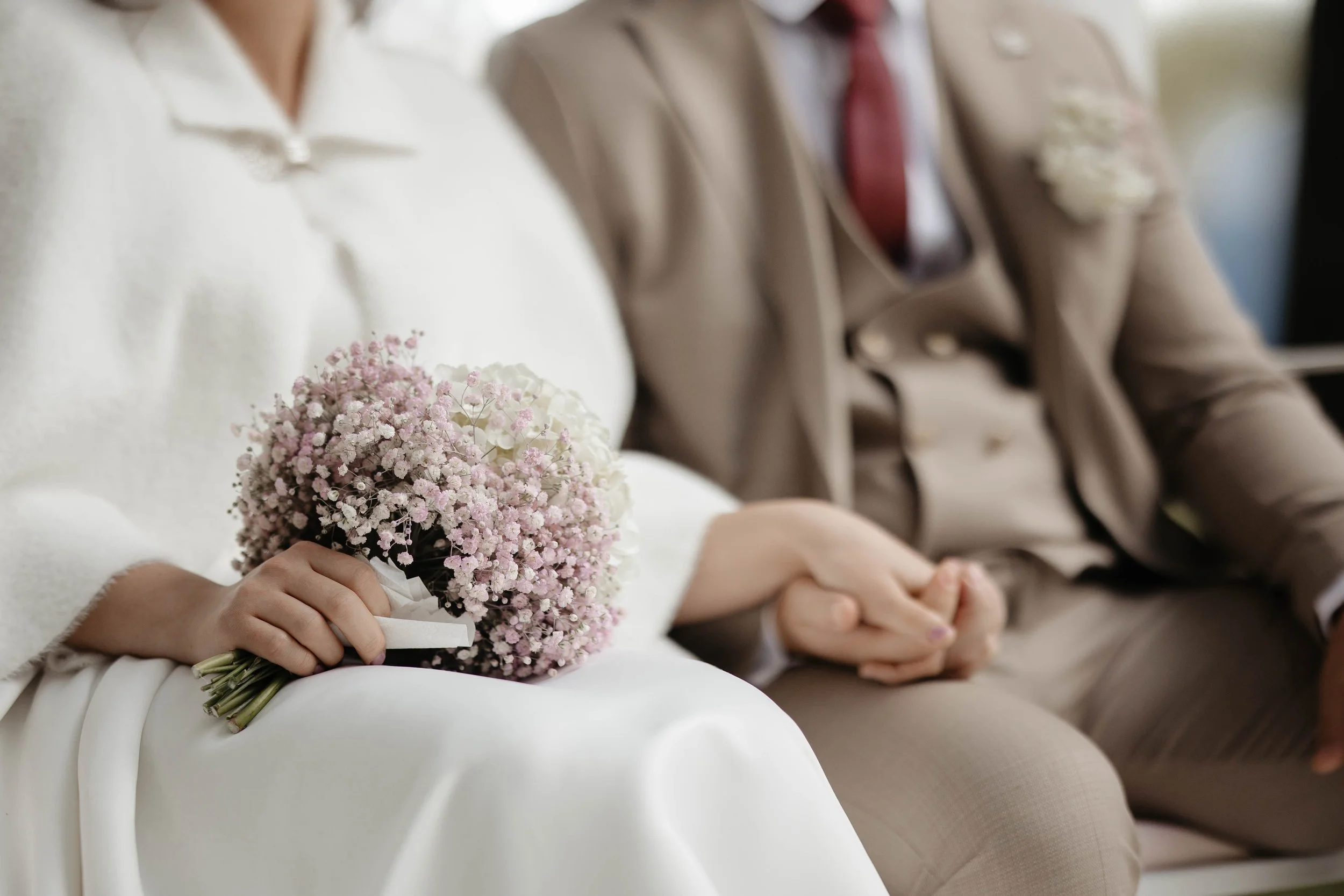 A close-up of a bride holding a bouquet of pink and white flowers, sitting next to a groom in a beige suit with a red tie, both dressed in formal attire at a wedding ceremony.