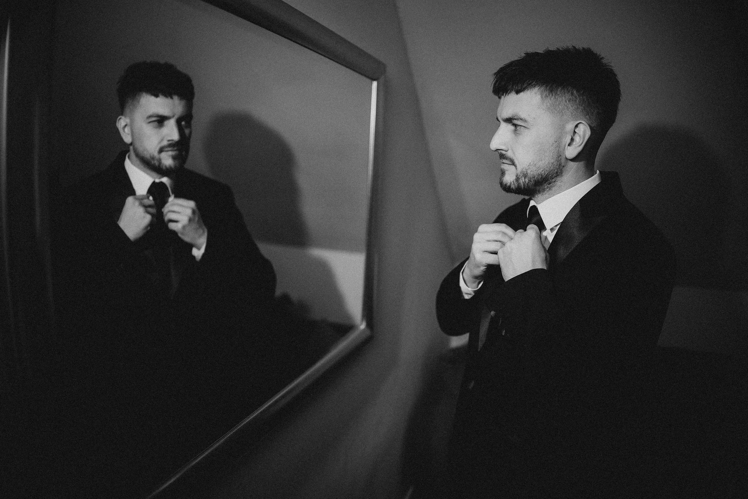 A man in a tuxedo adjusting his bow tie, seen in the mirror, with his reflection also visible in the photograph.