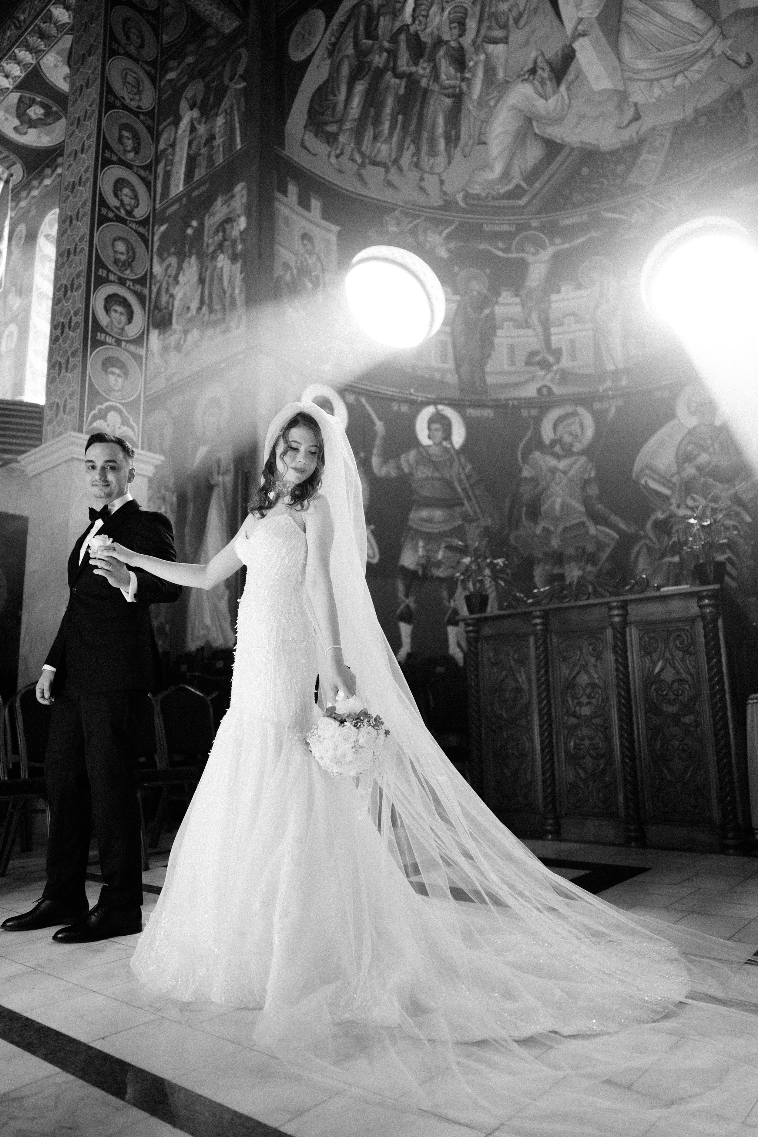 Black and white photo of a bride and groom in a church, with ornate religious murals and icons on the walls, sunlight streaming through circular windows.