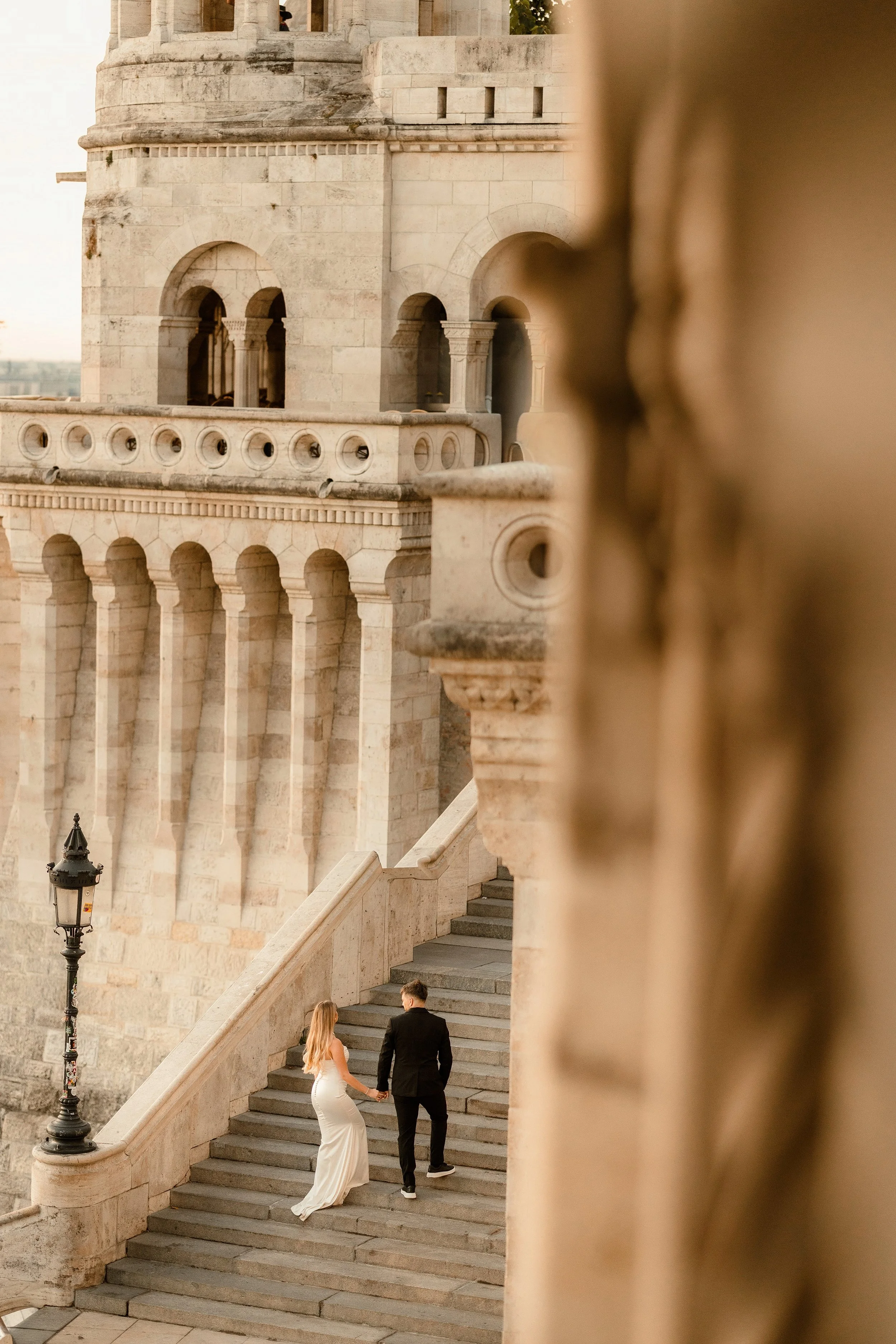 A bride and groom holding hands, walking down stone stairs outside of a historical building, with a street lamp nearby, during daylight hours.
