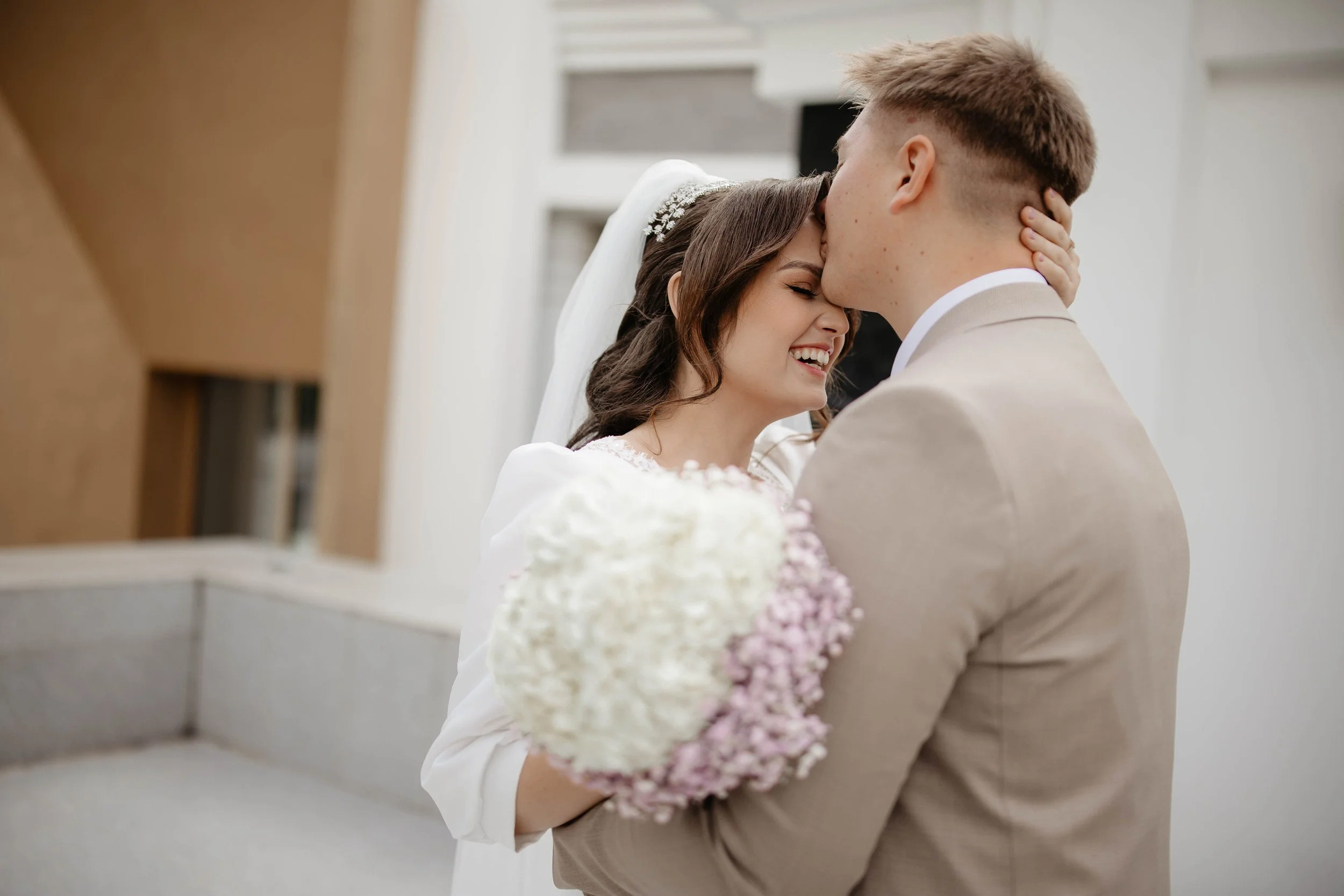 A bride and groom sharing a moment of intimacy, with the bride smiling and holding a bouquet of white and pink flowers, the groom gently touching the bride's head, both dressed in wedding attire inside a bright room.
