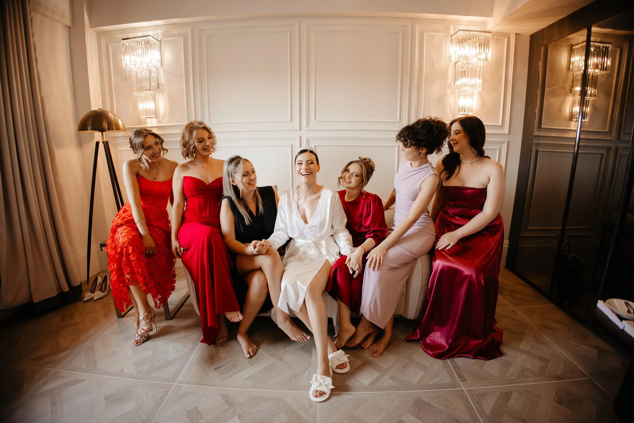 Group of seven women sitting on a beige couch in a stylish, well-lit room, with one woman in a white robe smiling surrounded by six women in red and pastel robes, some with heels, others barefoot.