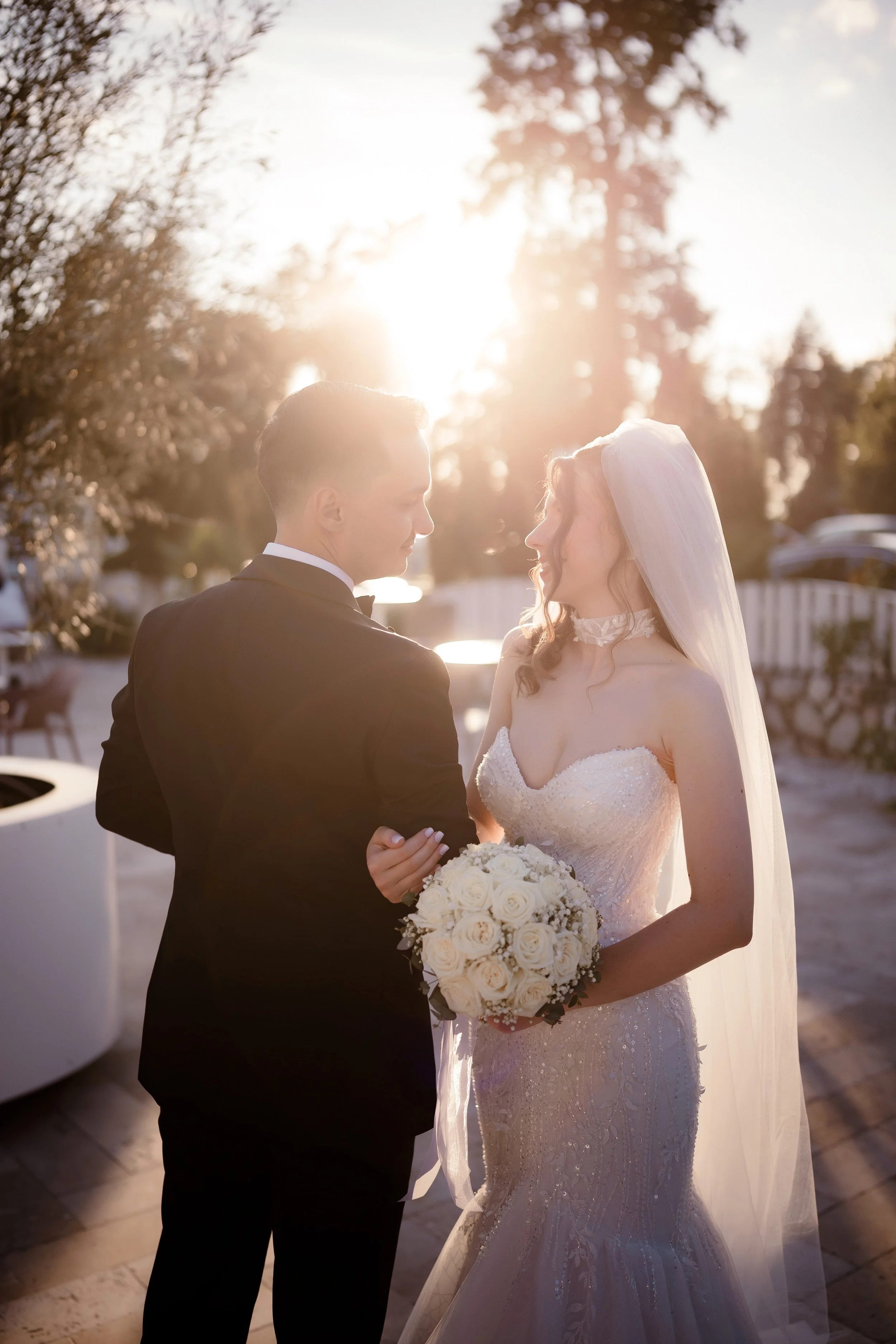 A bride and groom standing close together outdoors at sunset, facing each other, with the bride holding a bouquet of white roses.