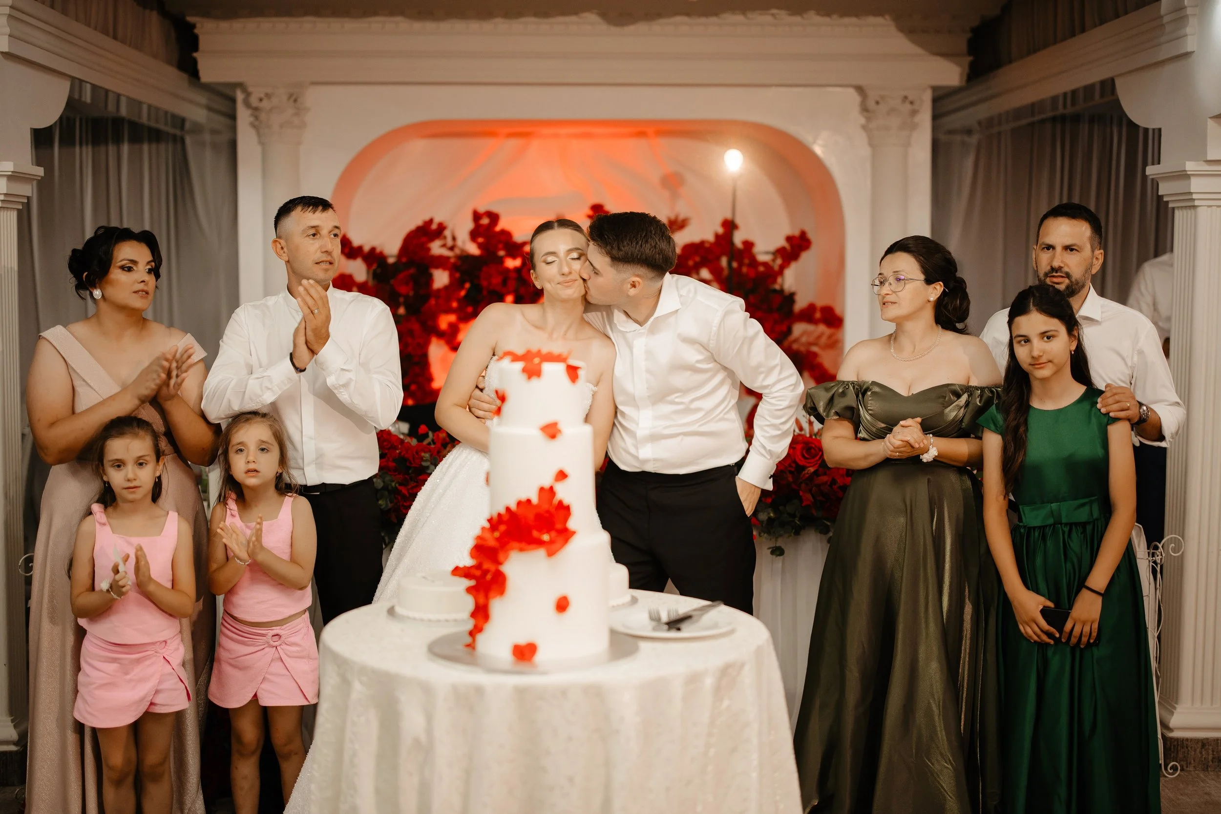 A group of people celebrating a wedding reception, with a bride and groom sharing a kiss in front of a wedding cake decorated with red flowers. Guests, including women and children, stand around clapping and smiling.