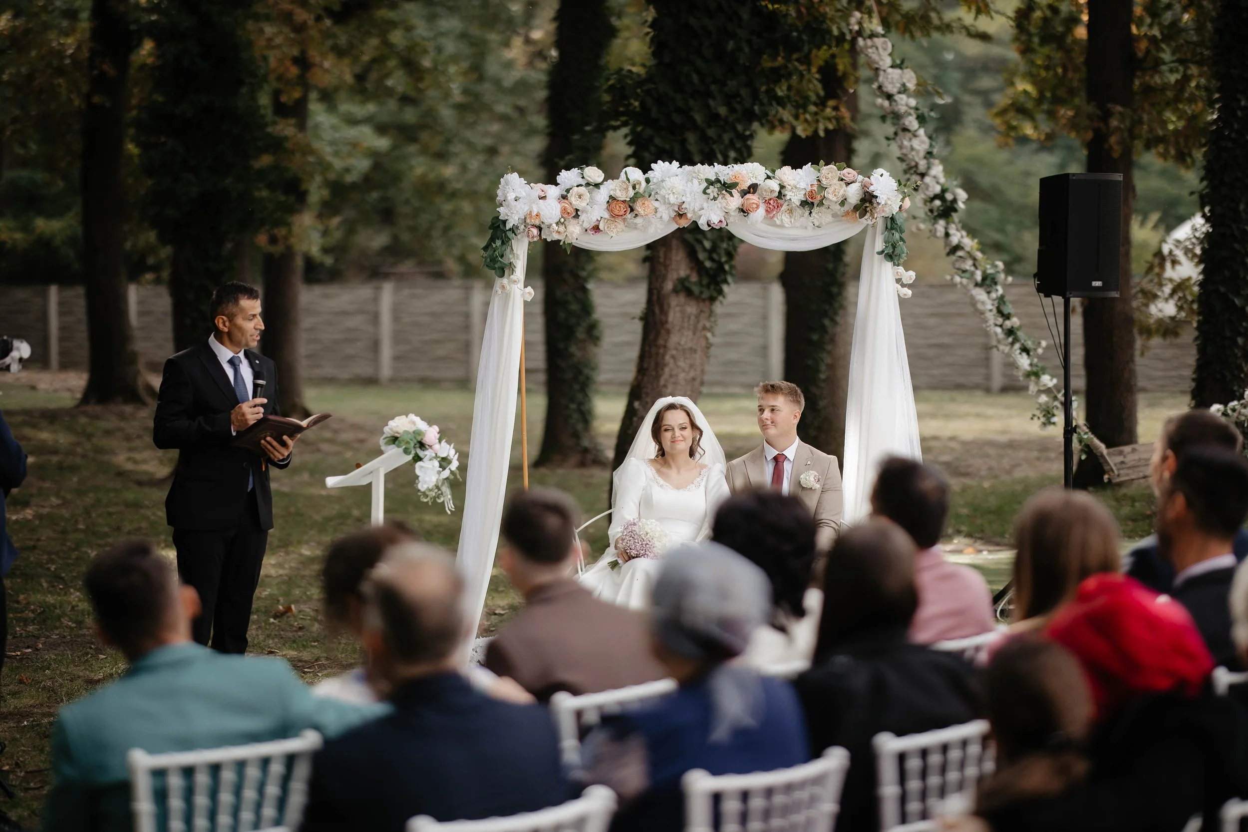 A couple getting married outdoors under a decorated floral arch, with an officiant speaking in front of them. Guests seated in white chairs are watching. Trees surround the scene.