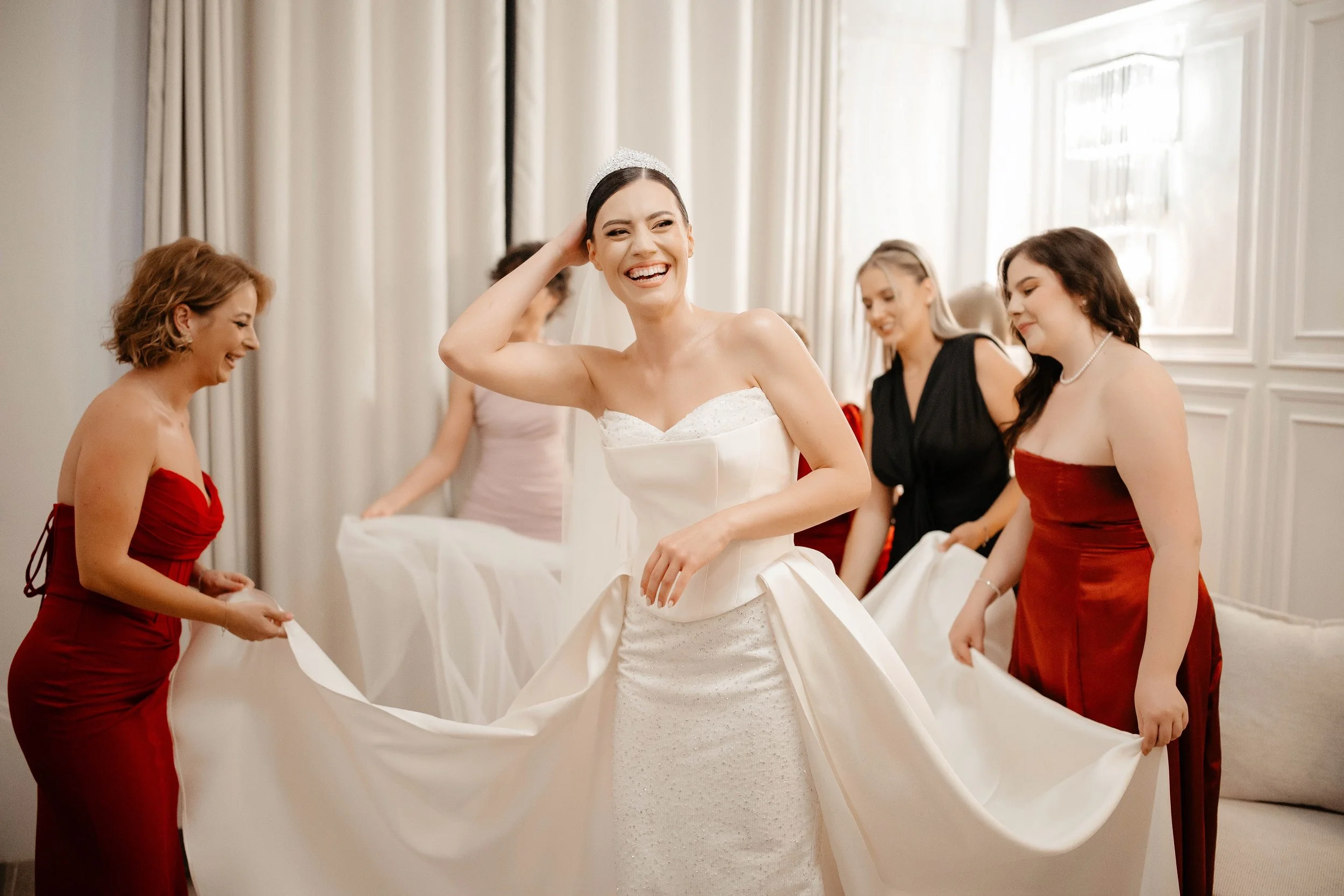 A bride in a white wedding dress smiling as she gets dressed with the help of women around her, some of whom are holding her dress and wearing red and black dresses.