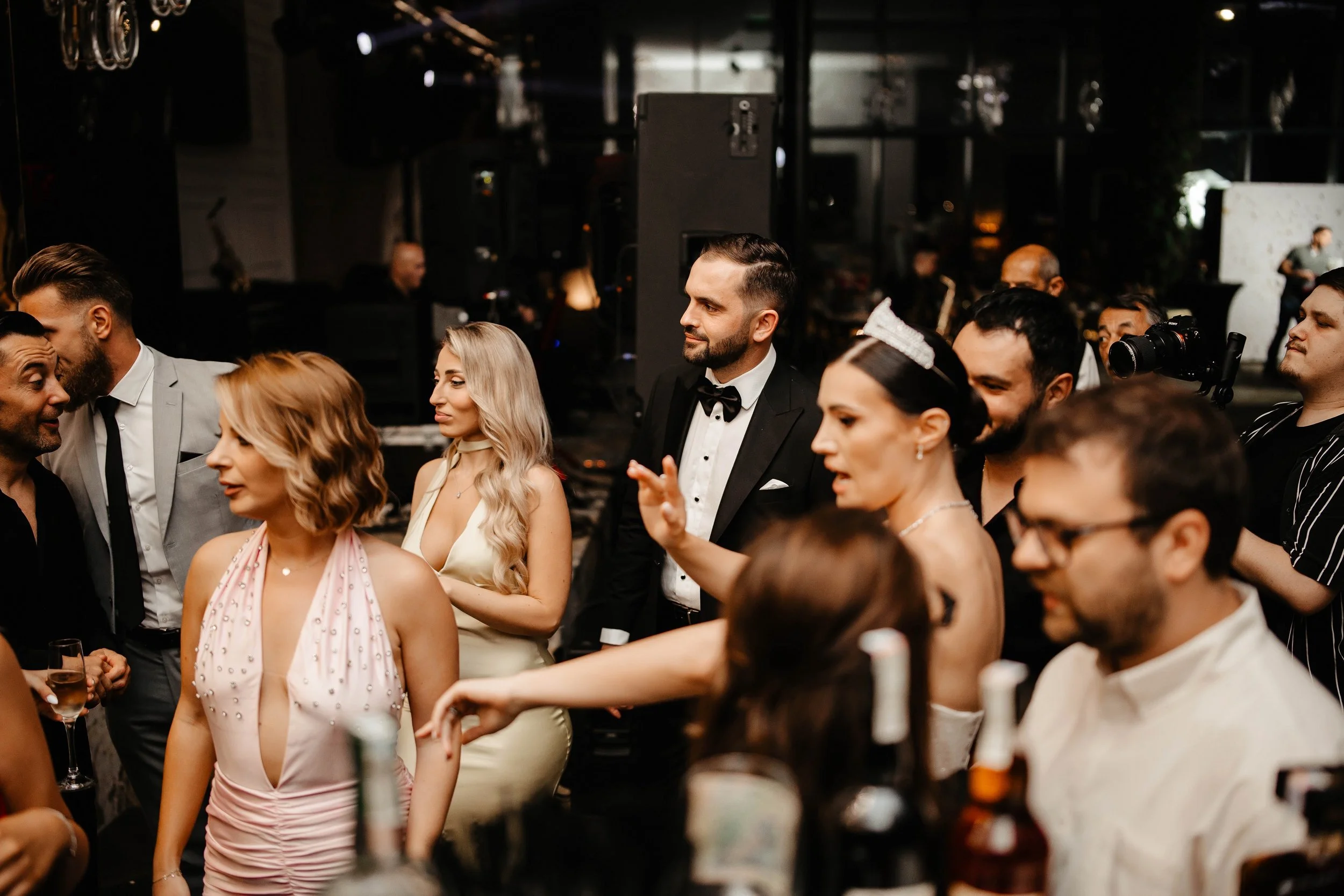 Group of people dressed in formal attire at a social event or party, engaging in conversations in a dimly lit indoor setting.