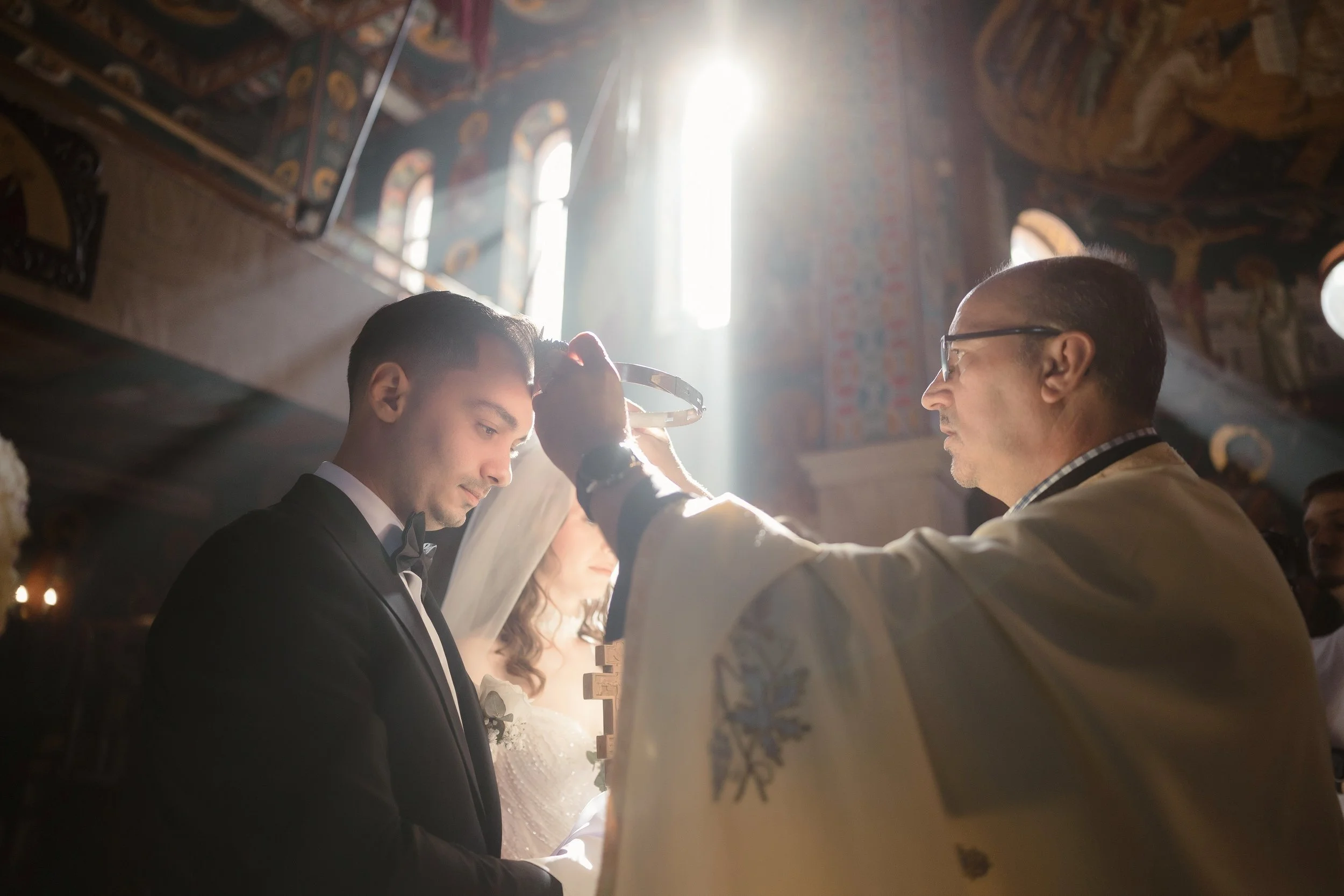 A wedding ceremony inside a church with sunlight streaming through a window, where a priest is placing a crown or similar item on the groom's head while the bride, groom, and officiant are present.