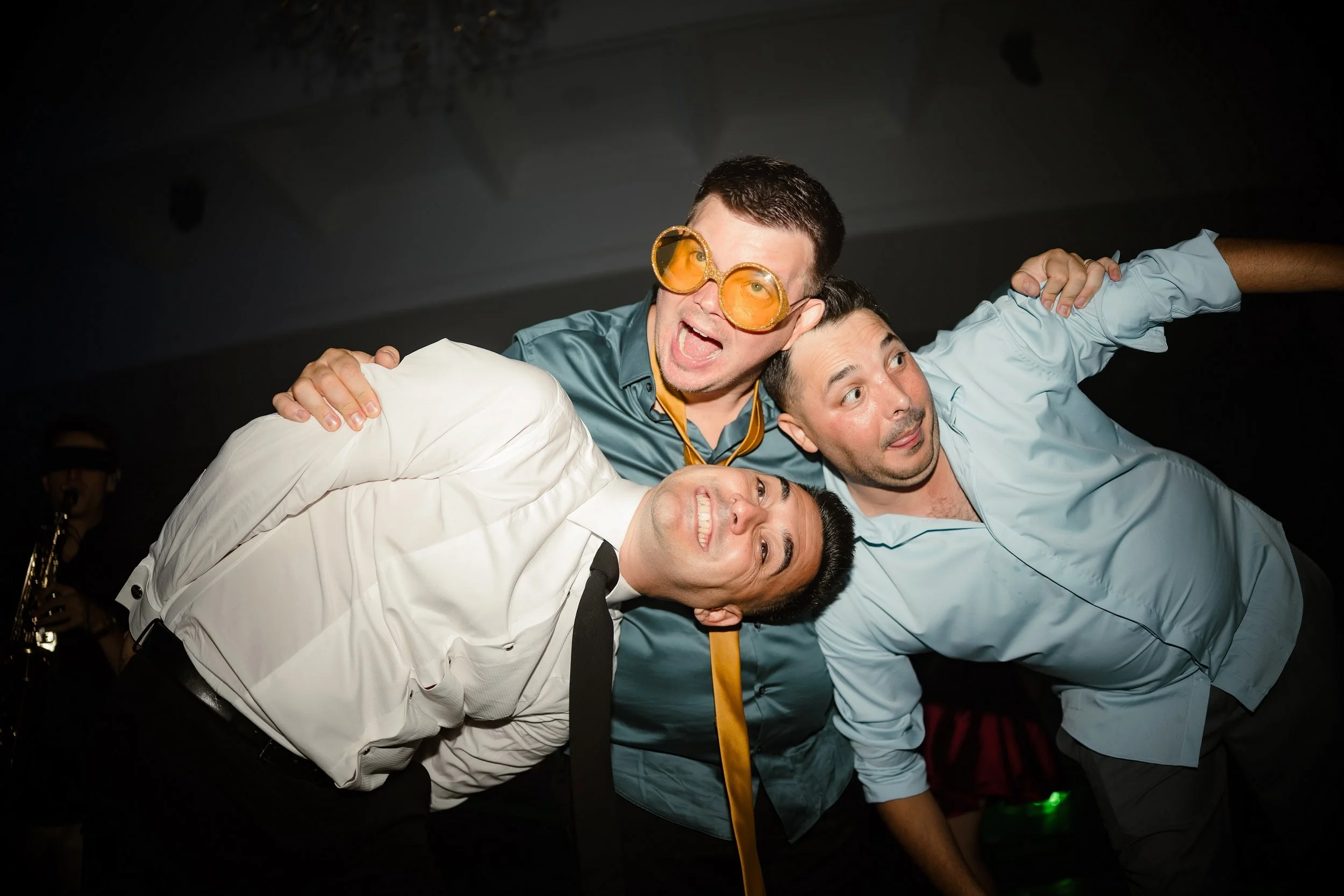 Four people at a party, two men and two women, are posing for a photo. The man in the center is wearing large orange sunglasses and a light blue shirt, and is smiling widely. The other three people are leaning in and smiling, with some of their hands
