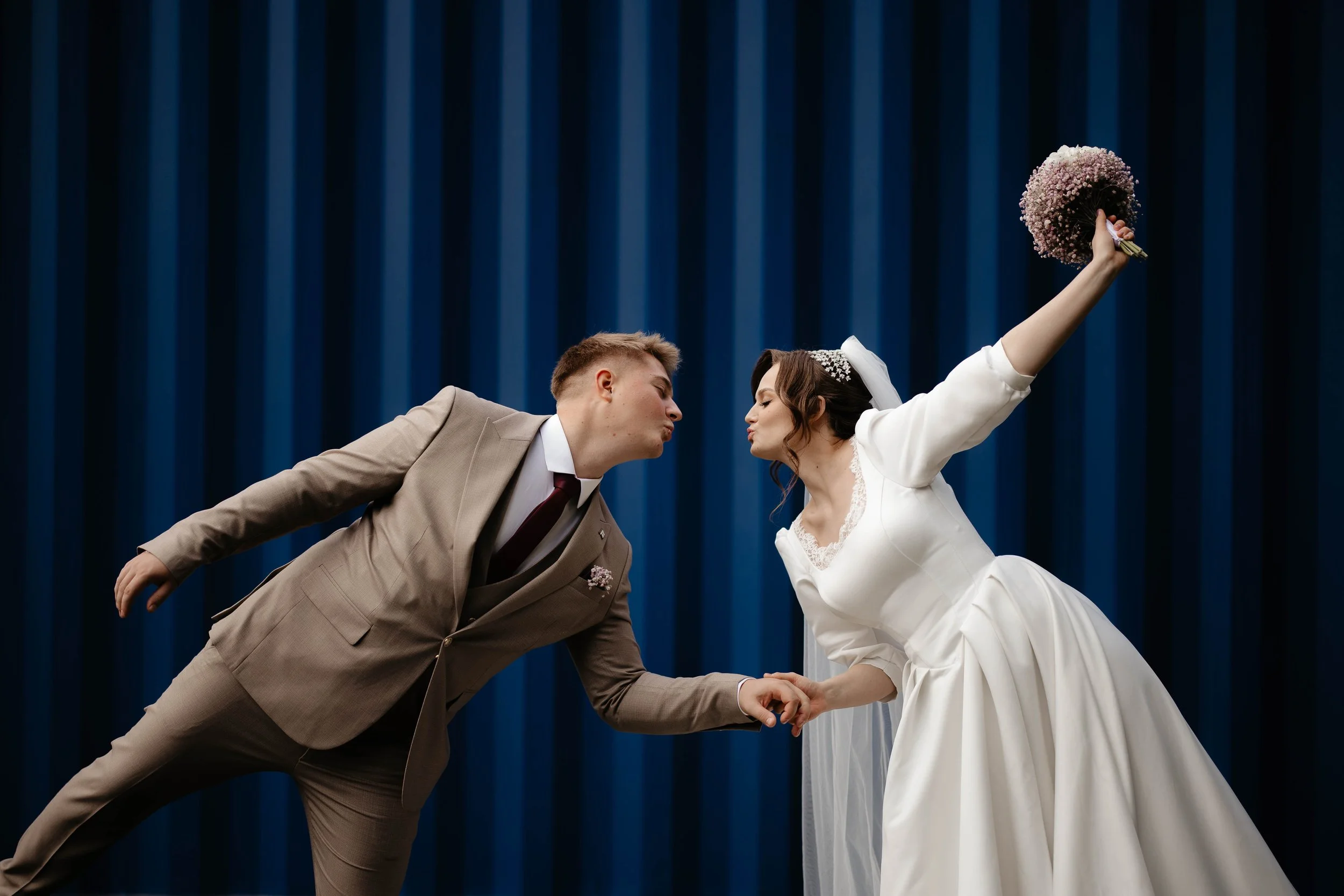 A bride and groom dance on a stage with a blue curtain background, touching foreheads and leaning towards each other while holding hands.