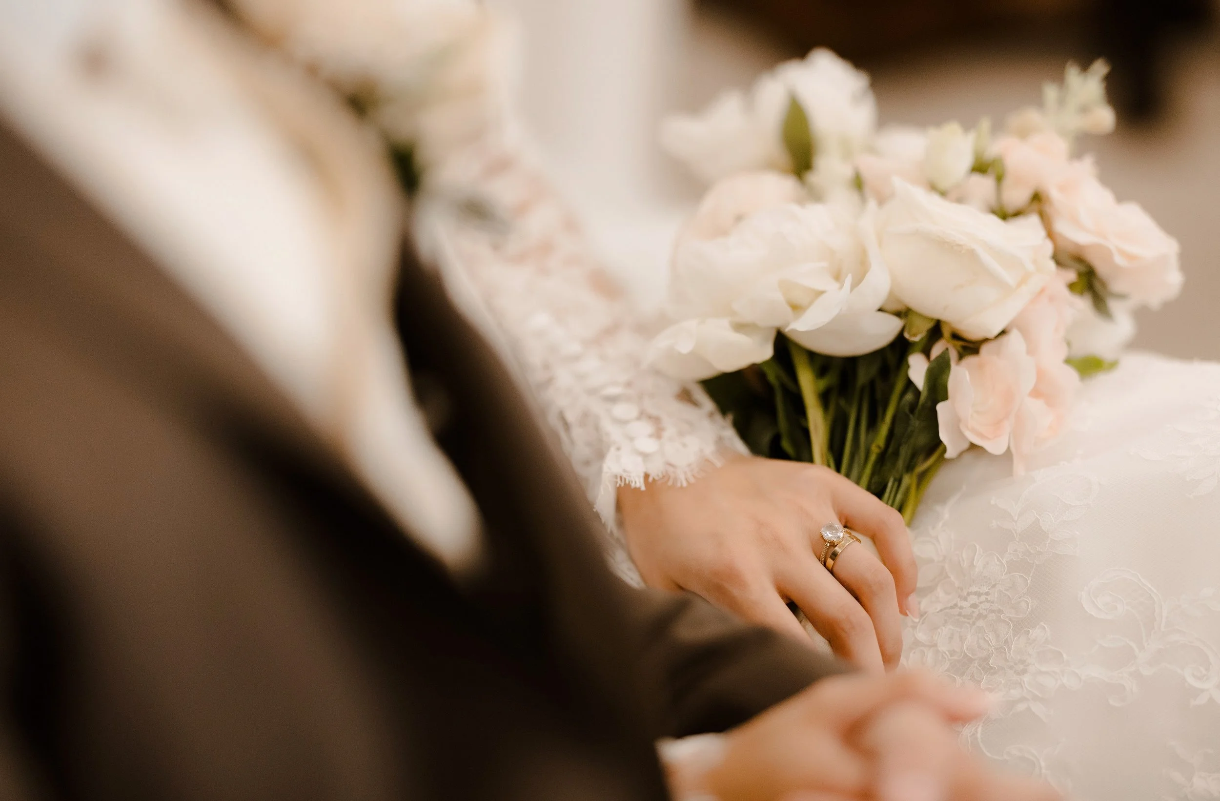 Close-up of a bride holding a bouquet of white and light pink flowers, wearing a lace wedding dress, with her hand showing a gold wedding ring. The image is slightly blurry and out of focus.
