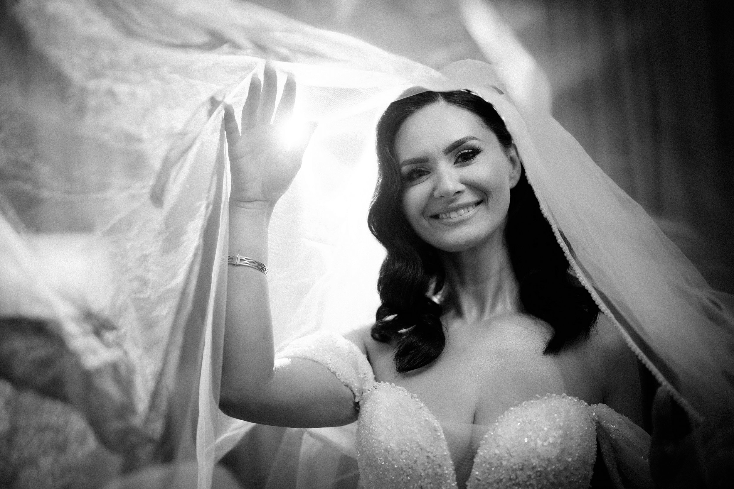 Black-and-white photo of a bride smiling, with long dark hair, in a wedding gown with lace details, holding a sheer veil above her head, and standing in front of a dark background.