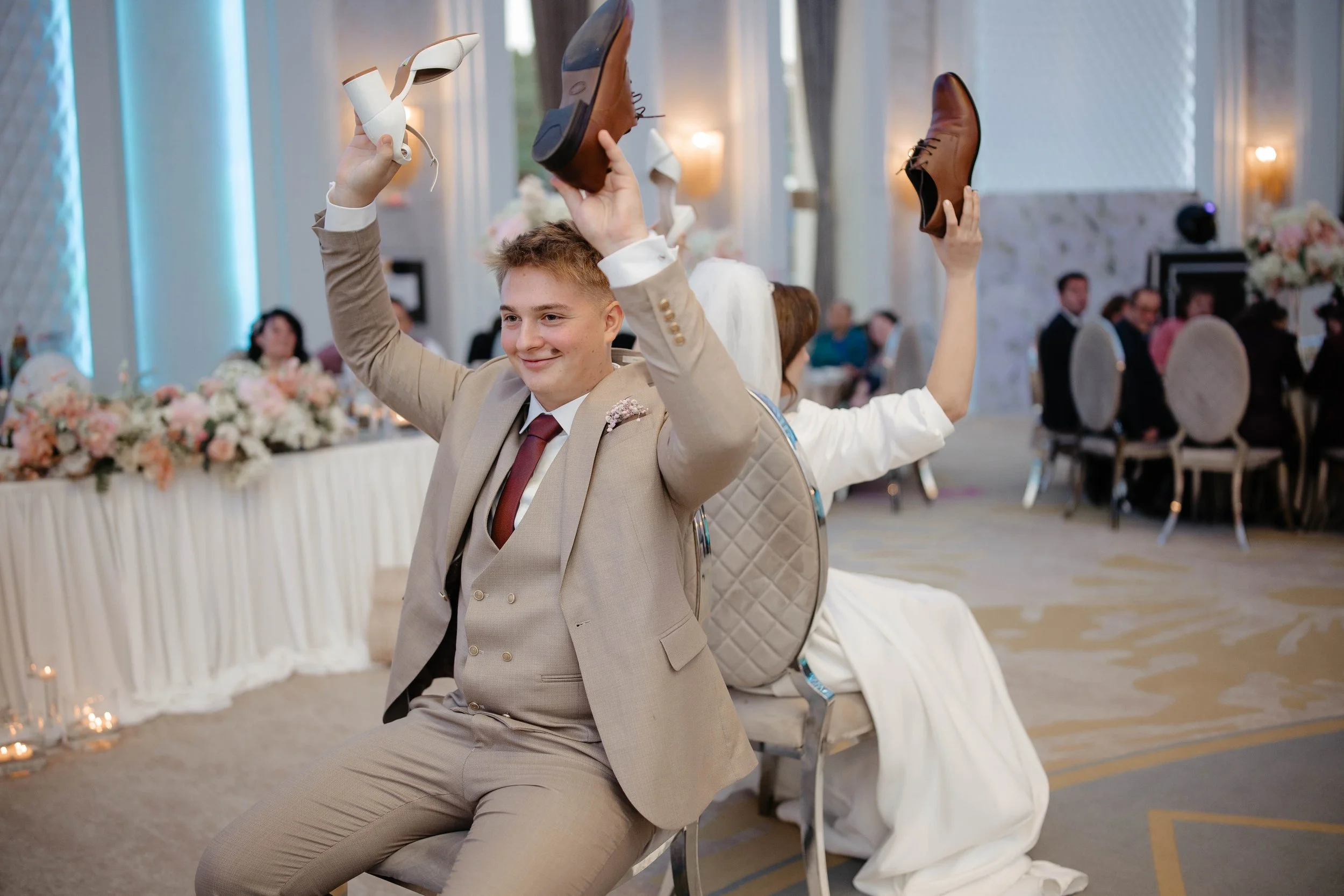 A young man in a beige suit is sitting in a banquet hall, playing a game with others in the background. He is holding a shoe in each hand, with a smile on his face. The hall is decorated with floral arrangements and multiple guests are seated at tabl