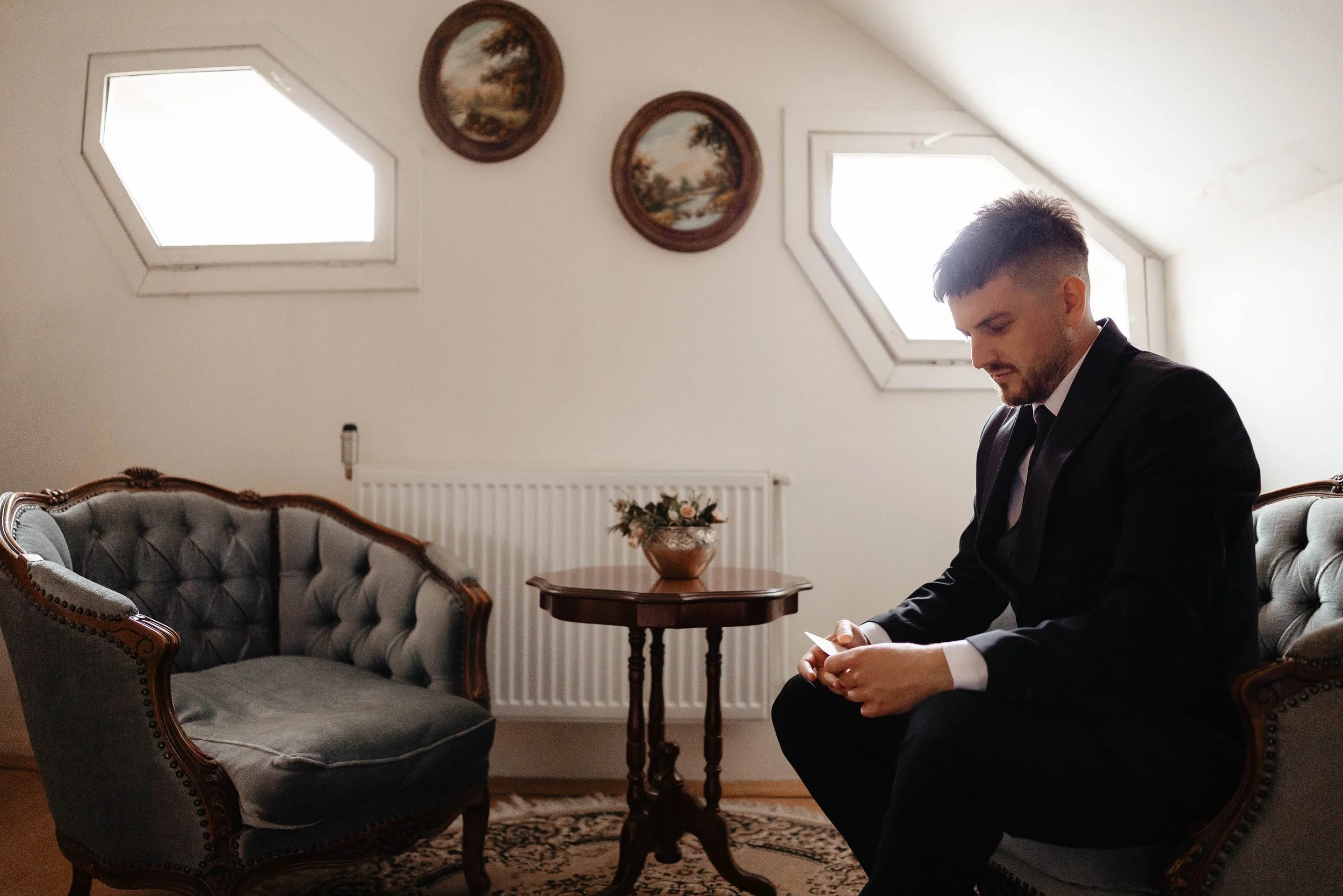 A man in a suit sitting on a vintage-style sofa in a room with two hexagonal windows and two landscape paintings on the wall. There is a small round table with a vase of flowers in front of him.