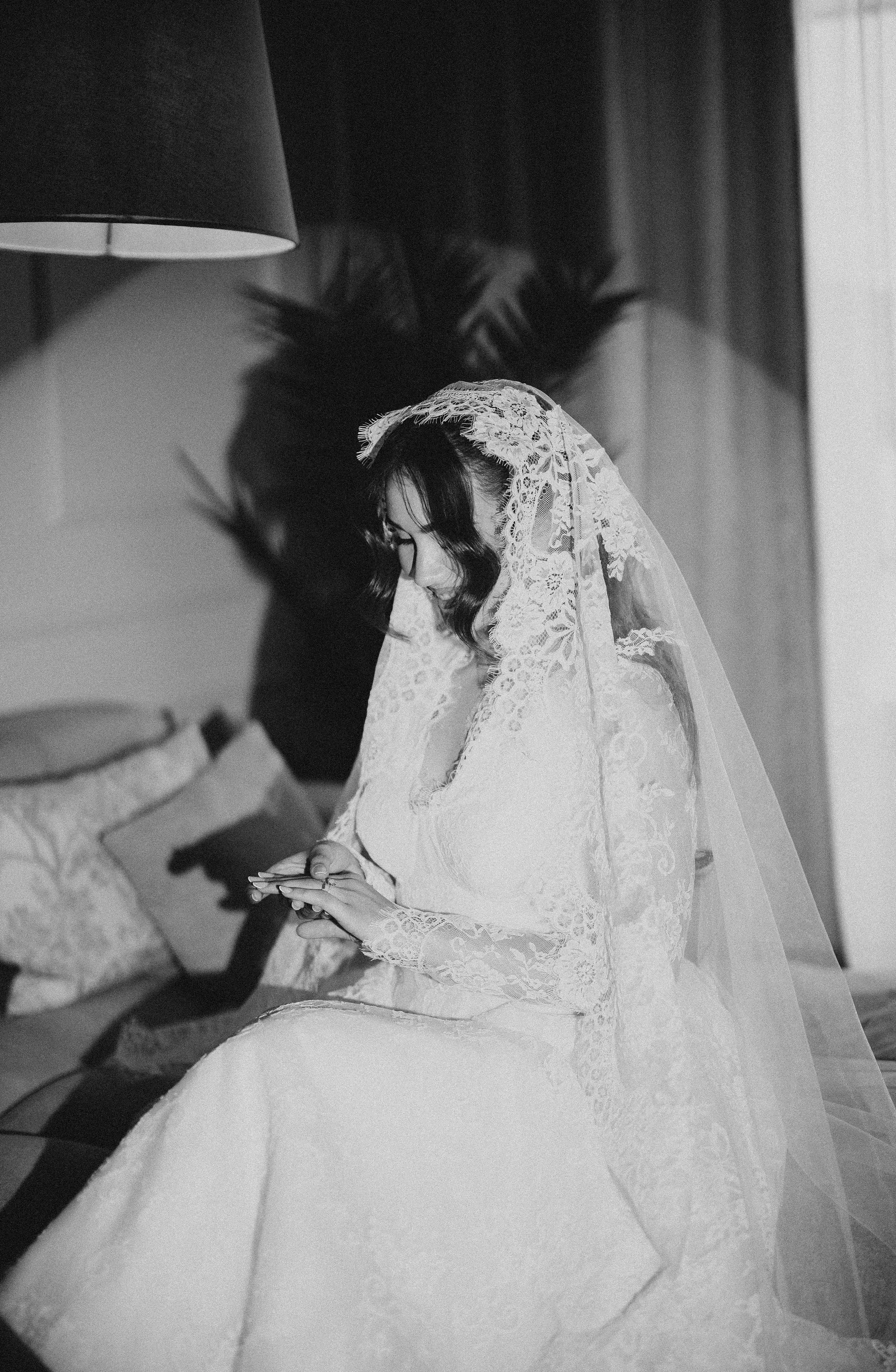 A bride in a lace wedding dress and veil sits on a bench, looking down and holding her phone, with her shadow cast on the wall behind her.