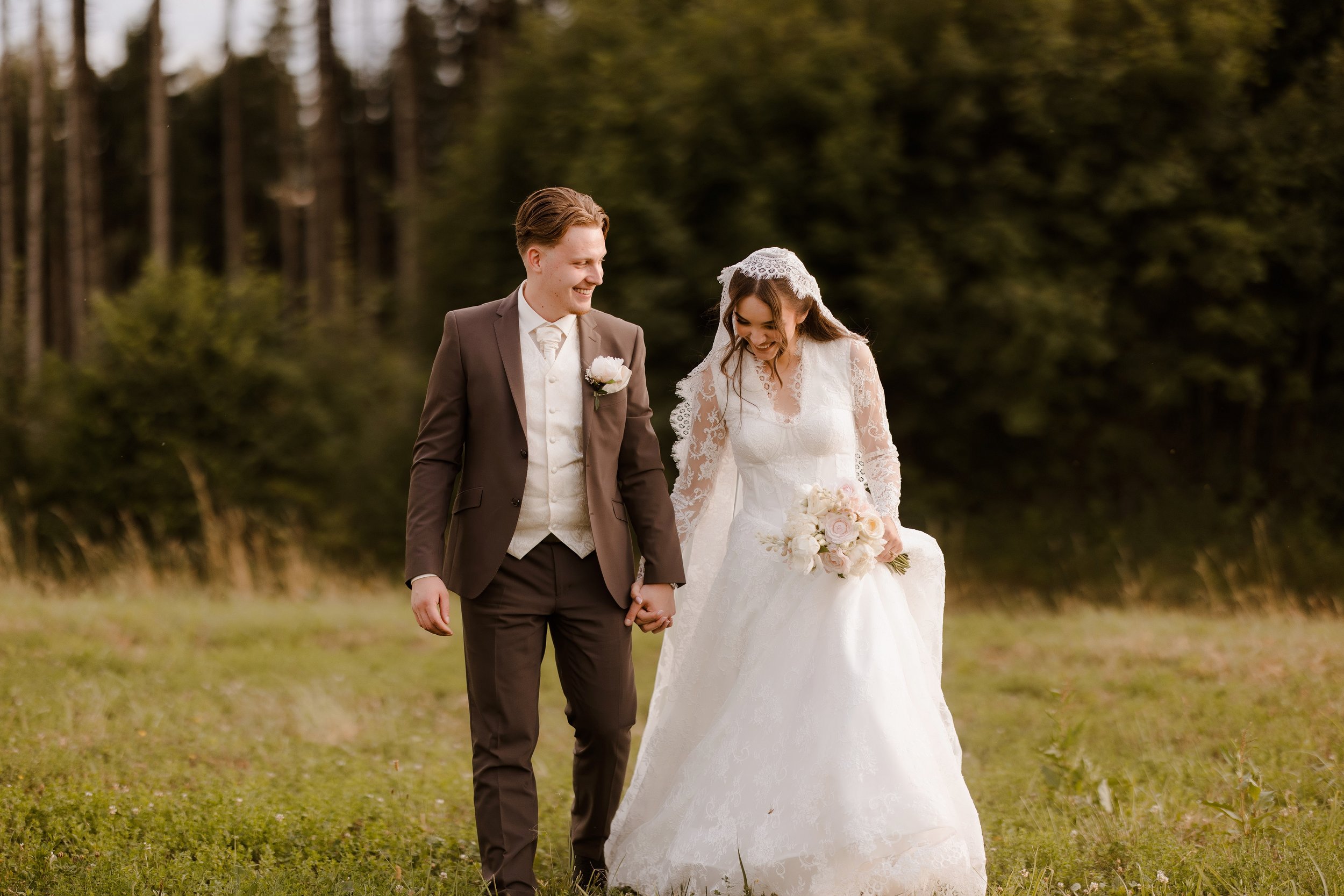 A bride and groom holding hands, walking outdoors on a grassy area with trees in the background. The bride is dressed in a white lace wedding gown with a veil, and the groom is in a brown suit with a cream vest and tie. They are smiling and looking a