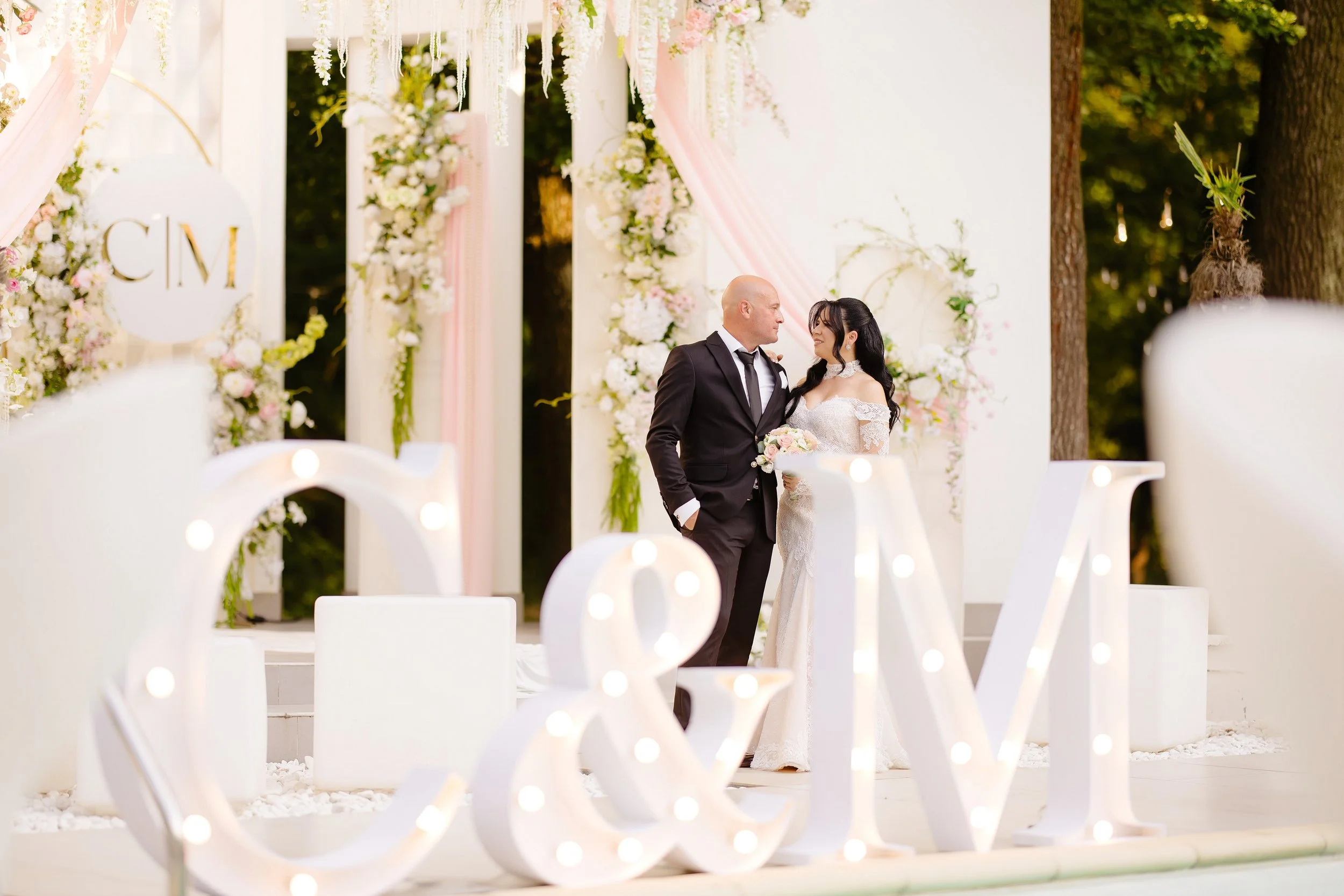 A bride and groom standing close together at their wedding ceremony outdoors, surrounded by floral decorations and greenery.