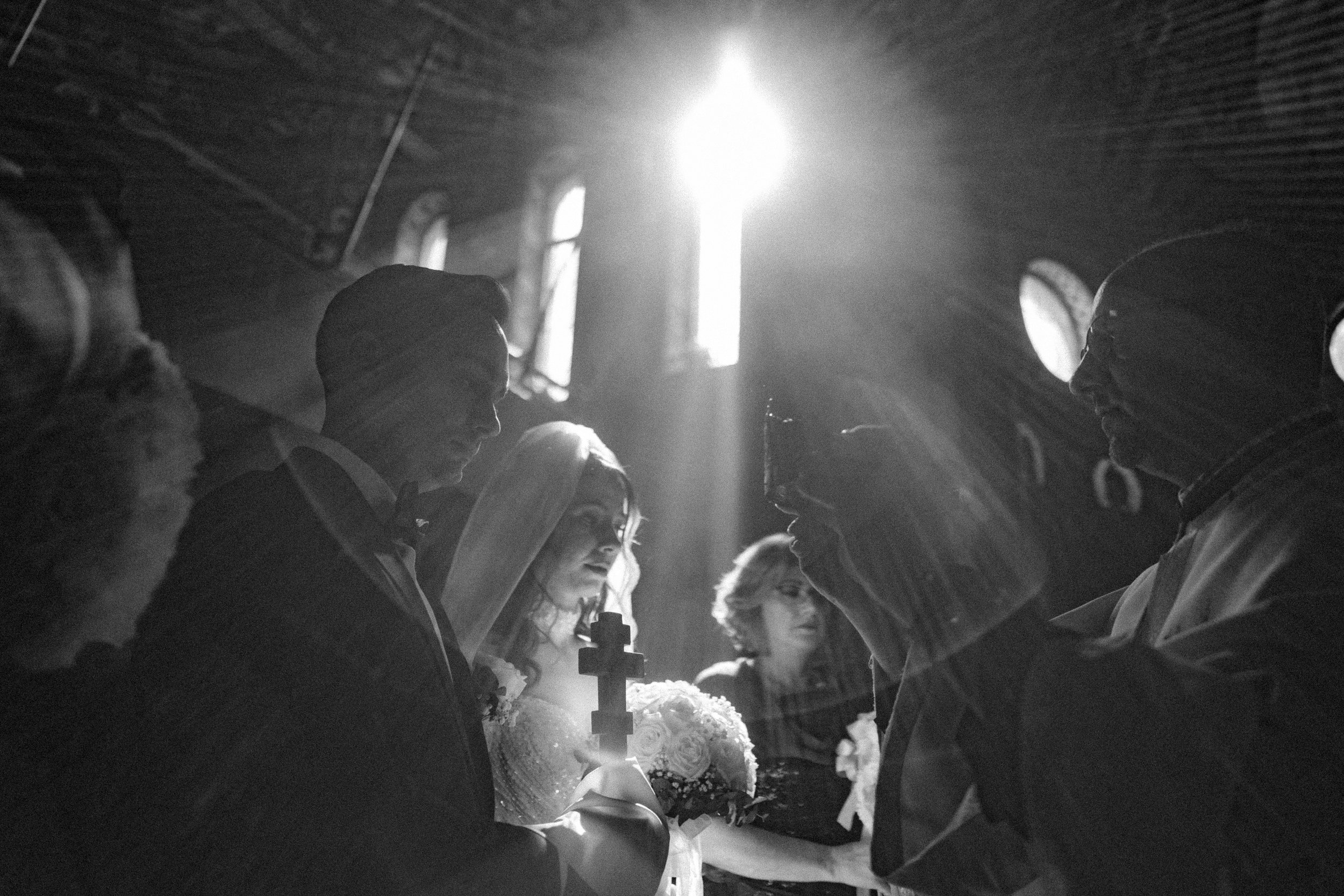 Black and white photo of a group of people holding candles and flowers in a solemn gathering inside a church. The sunlight streaming through windows highlights their faces and candles.