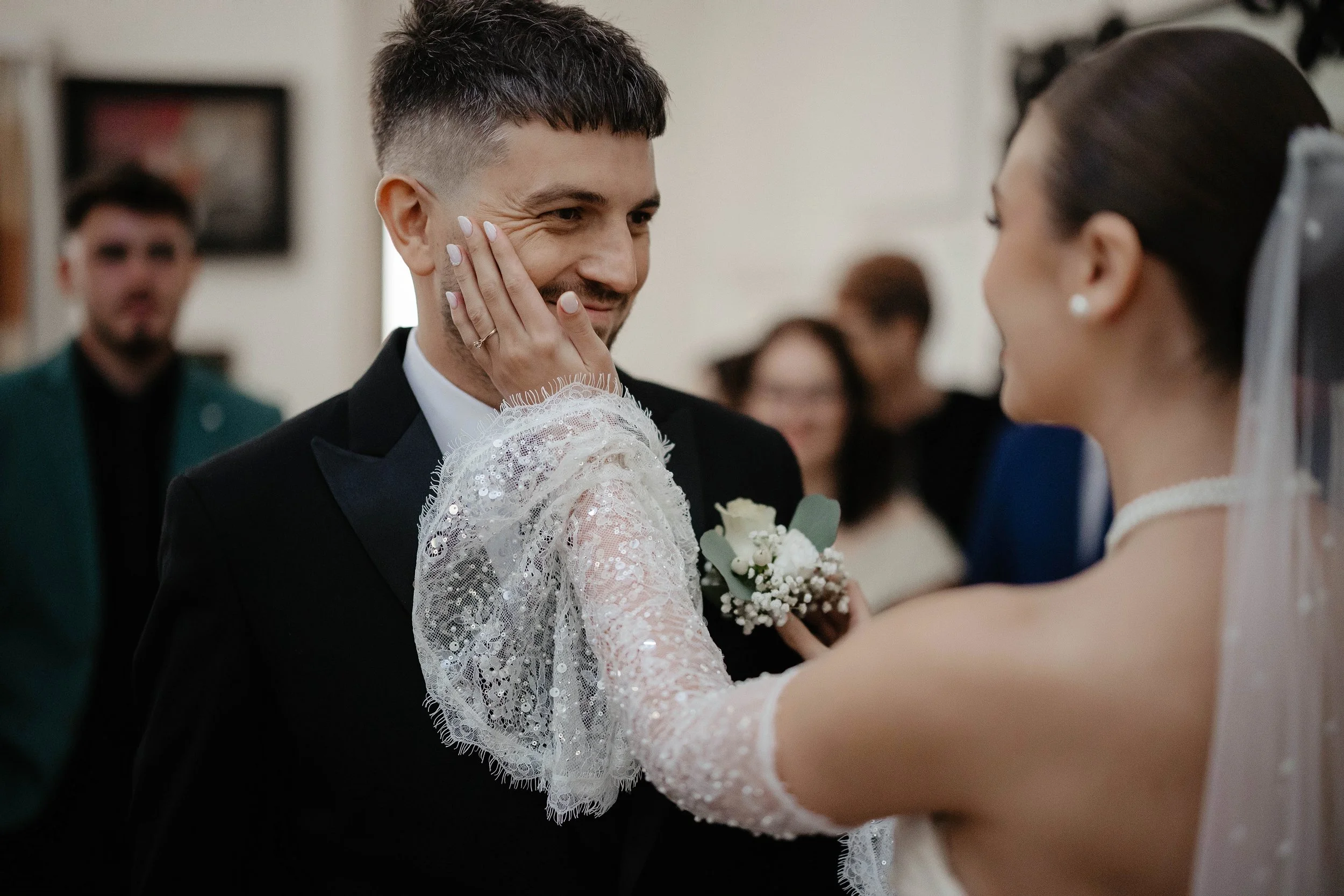 A couple is sharing a kiss at their wedding ceremony, with the groom smiling and the bride holding his face. The groom wears a black tuxedo and the bride has a white lace dress with long sleeves. In the background, a man in a dark suit stands among o