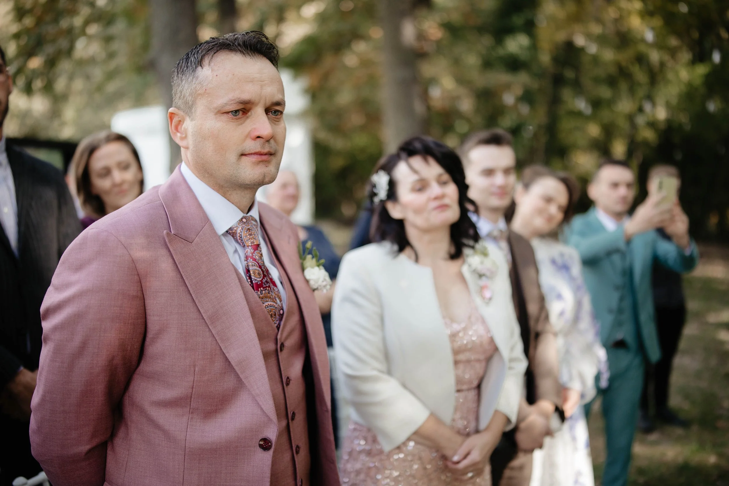 A group of people dressed in formal attire, standing outdoors with trees in the background, possibly at a wedding or ceremony.