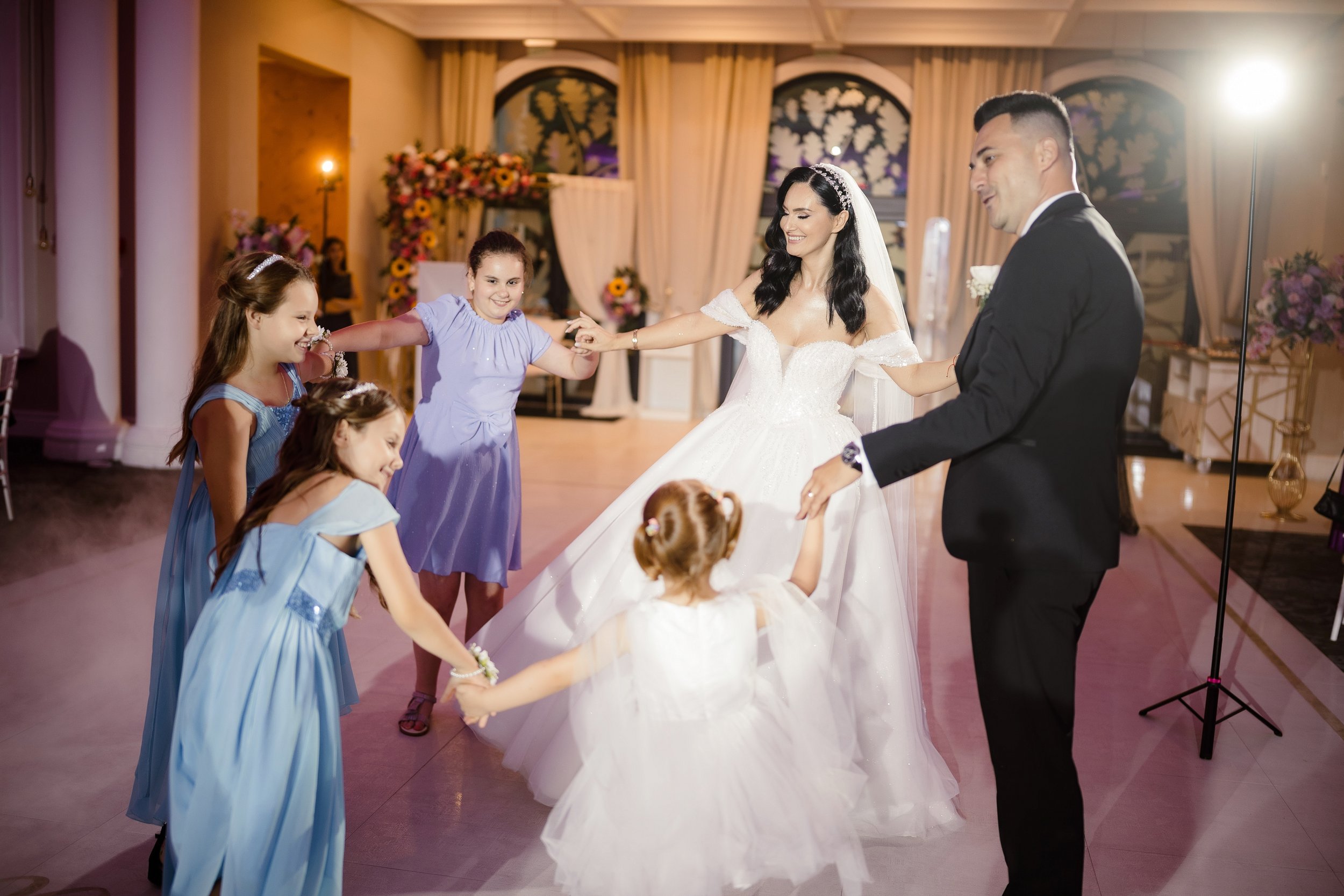 A bride and groom dancing with young girls at a wedding reception. The bride is in a white wedding gown, and the groom is in a black tuxedo. The girls are wearing pastel-colored dresses and smiling.