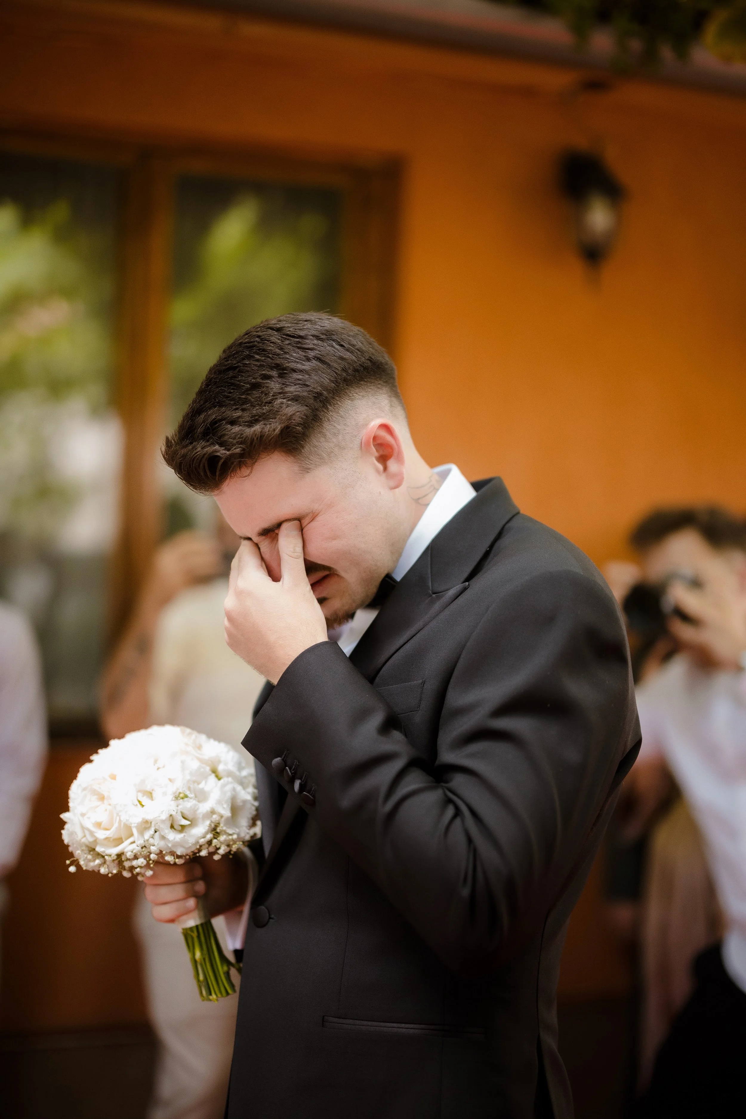 A man dressed in a black tuxedo, holding a bouquet of white flowers, with his eyes closed and hand covering his face in an emotional moment at a wedding reception.
