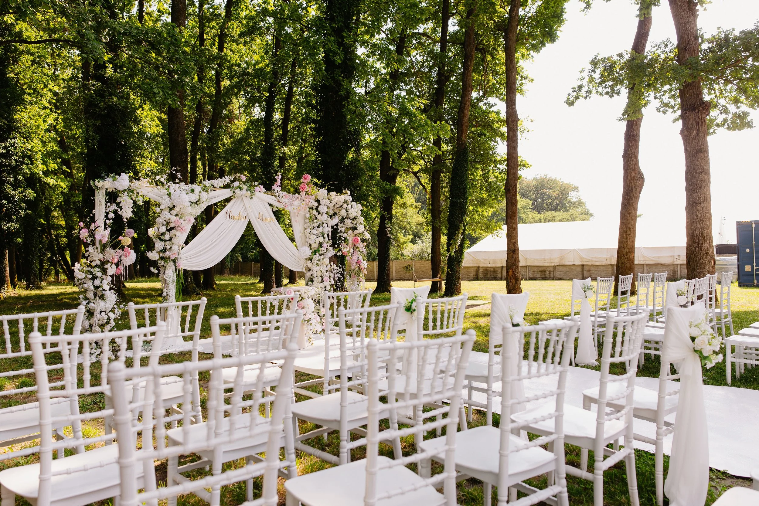 Outdoor wedding setup with white chairs and floral arch under a large tree, sunny day.