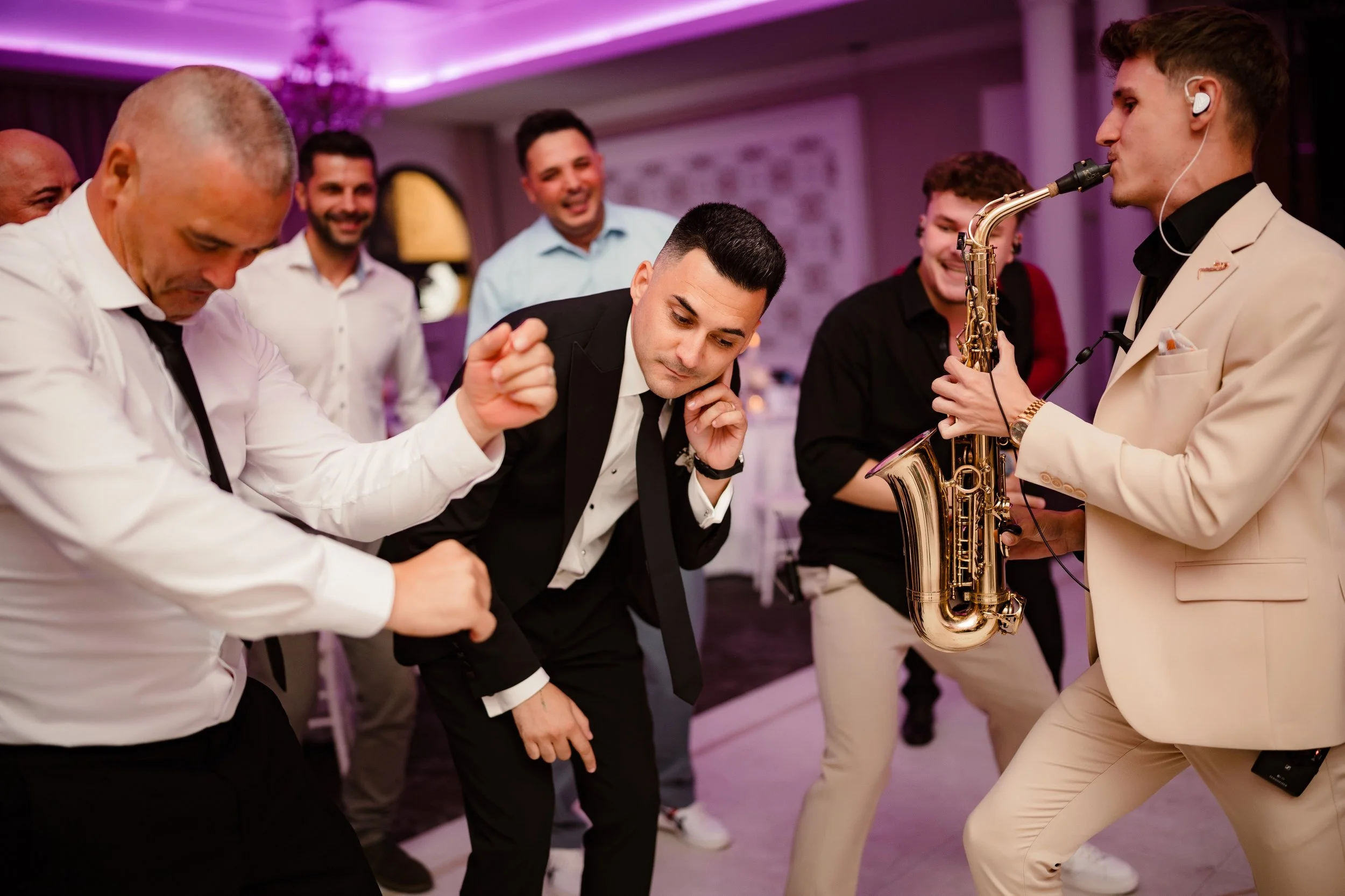 Men dancing and enjoying a lively party with saxophonist playing music, surrounded by smiling friends in an elegant indoor setting with pink and purple lighting.
