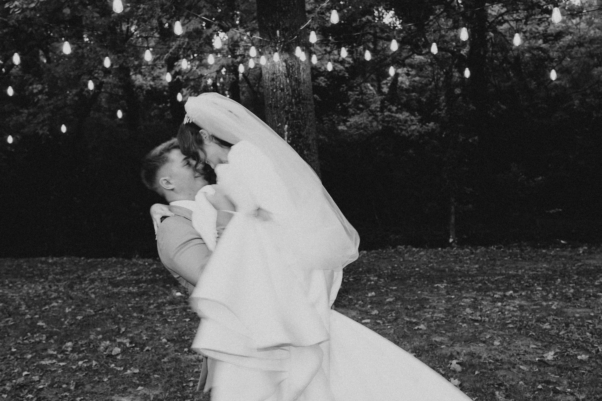 A couple dancing outdoors at night with string lights overhead, the woman in a white dress and the man in a light suit, surrounded by trees and fallen leaves.
