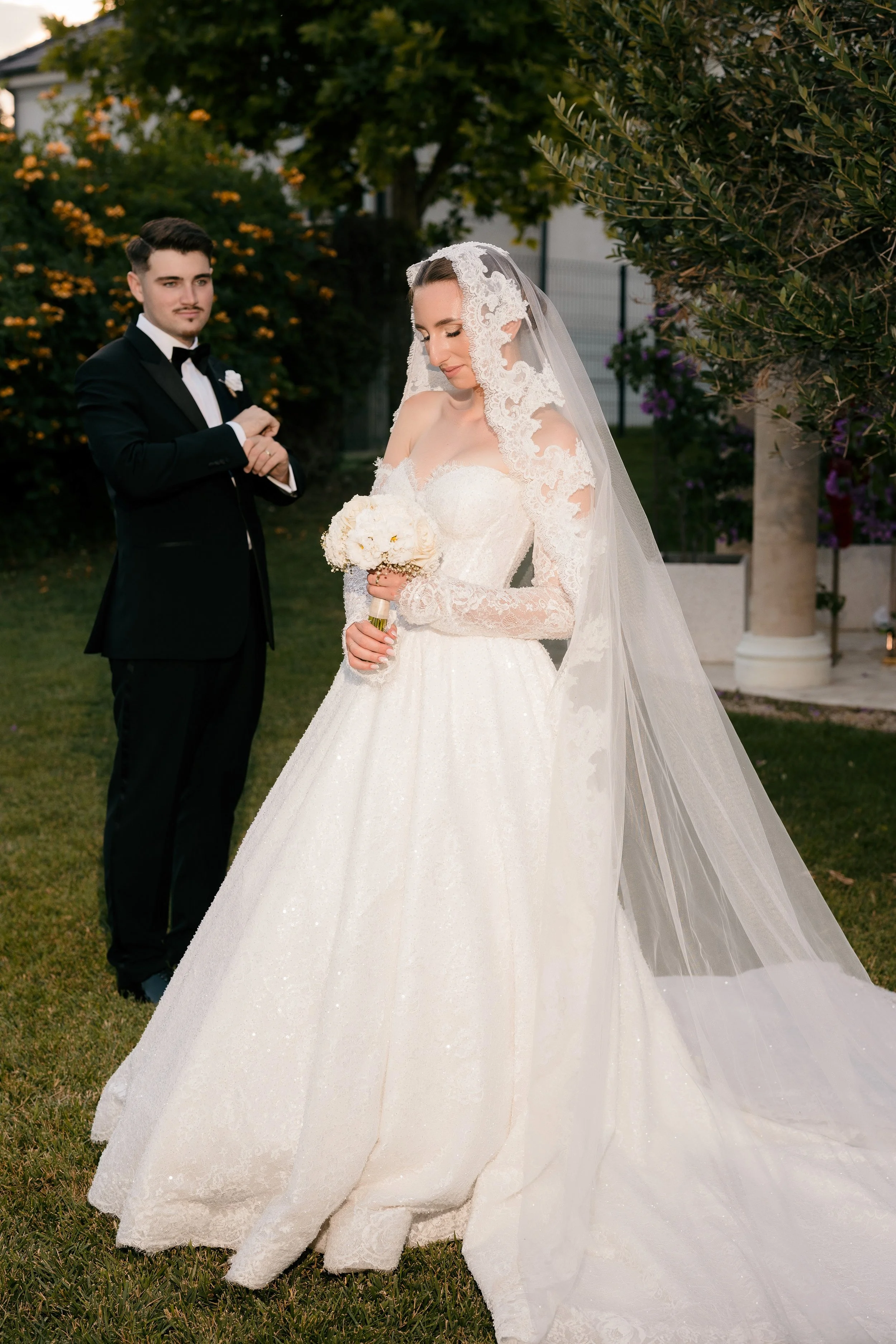 Bride in a lace wedding gown and veil holding a bouquet of white flowers, with a groom in a black tuxedo standing nearby, outdoors with trees and flowers in the background.