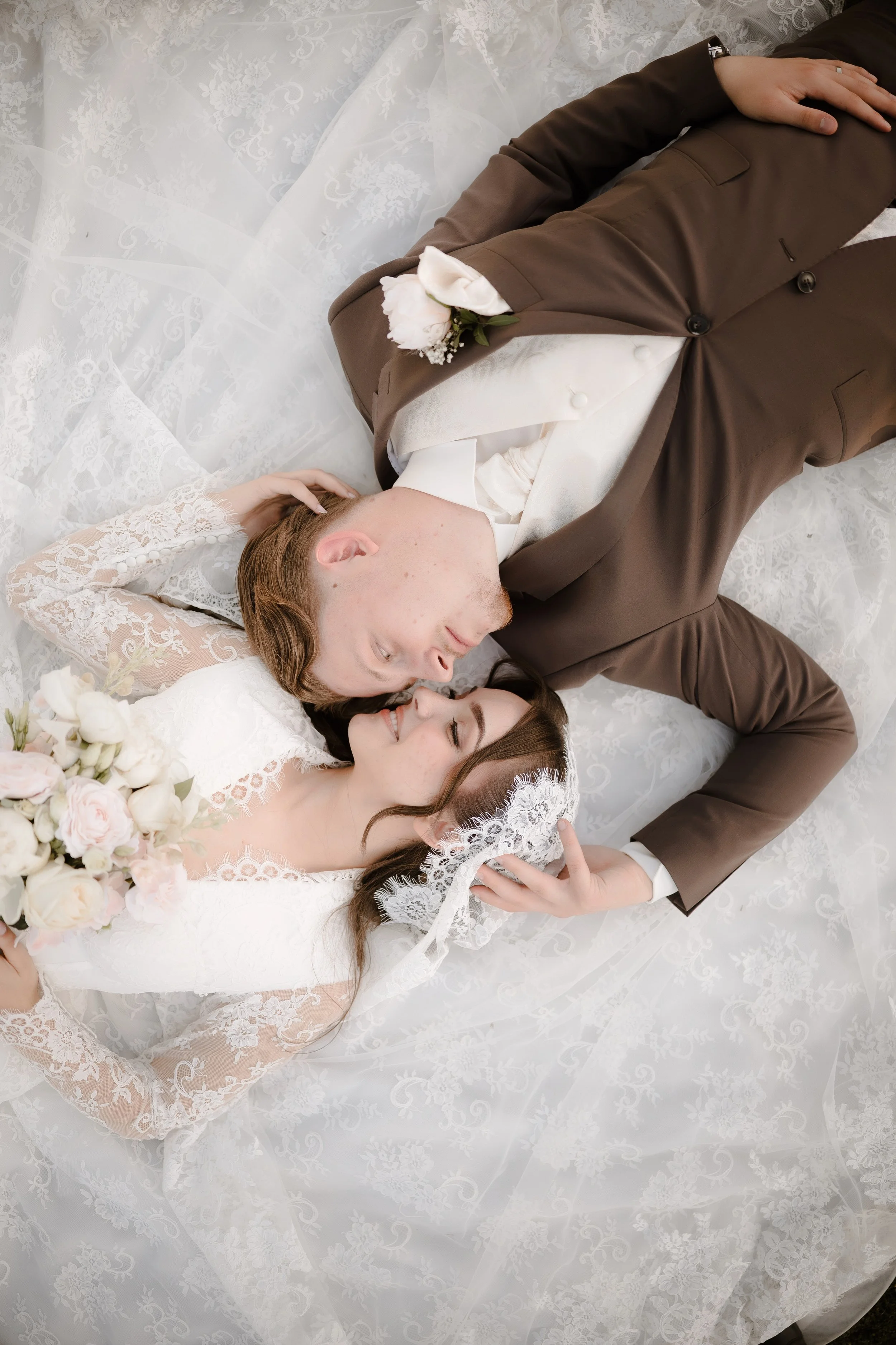 A couple dressed in wedding attire lying on a white lace-covered surface and touching foreheads, with their eyes closed and wearing wedding rings.
