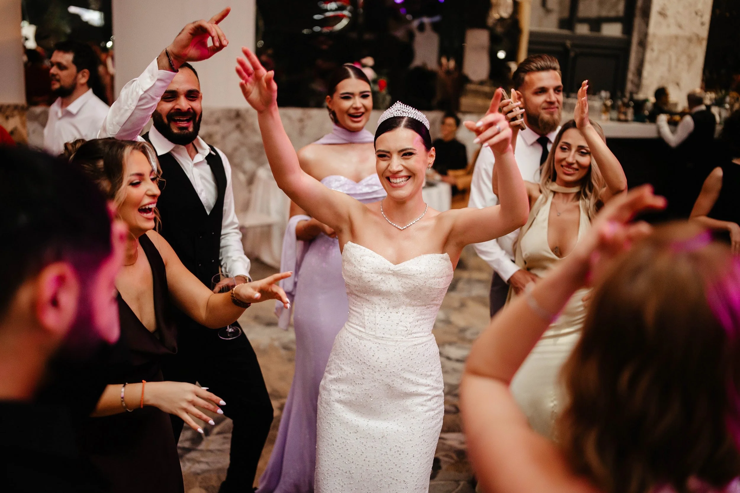 People dancing and celebrating at a wedding reception, with the bride wearing a white strapless gown and tiara, smiling and raising her arms.