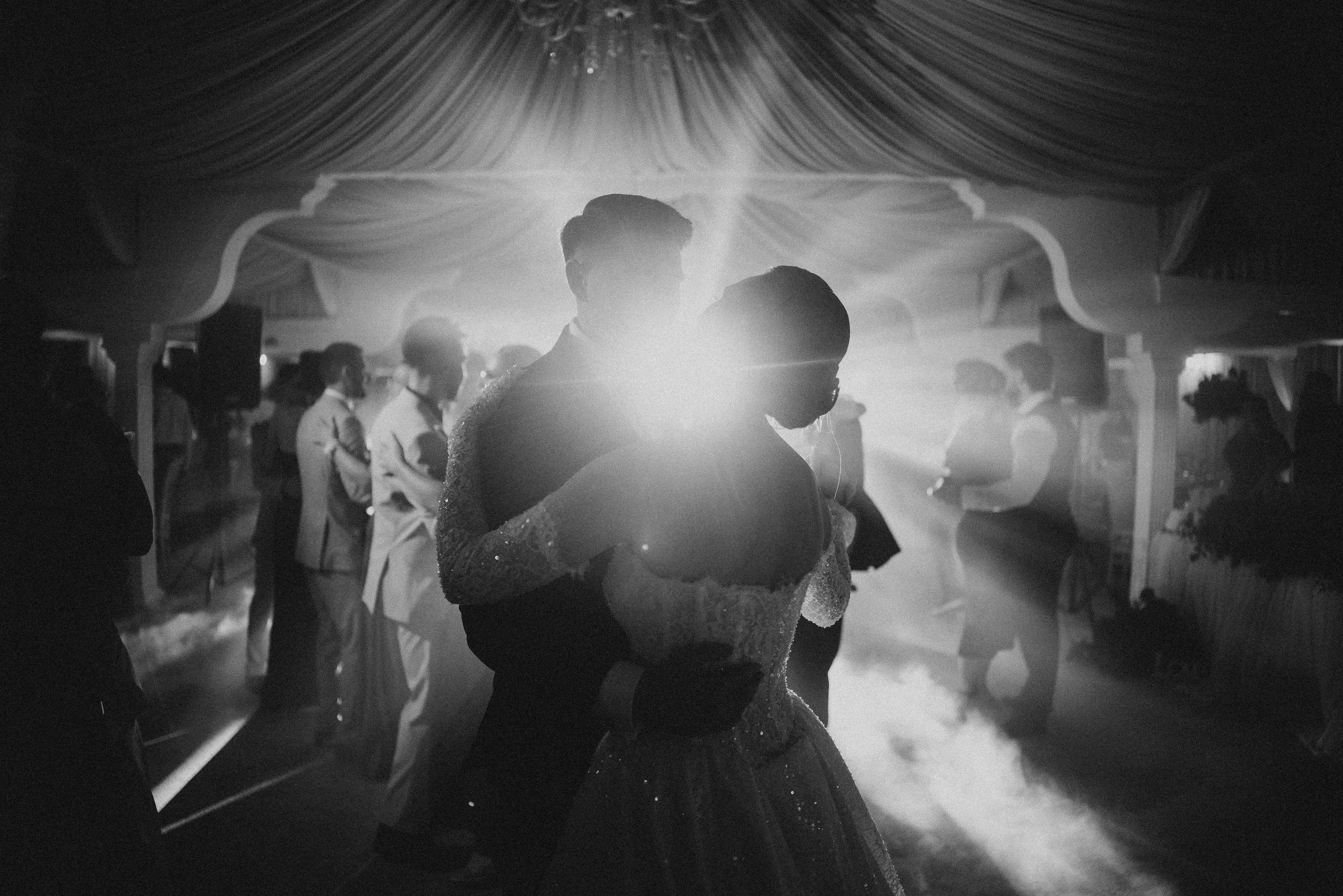Black and white photo of a bride and groom dancing closely at their wedding reception, surrounded by guests, with draped curtains overhead and lighting creating a romantic atmosphere.