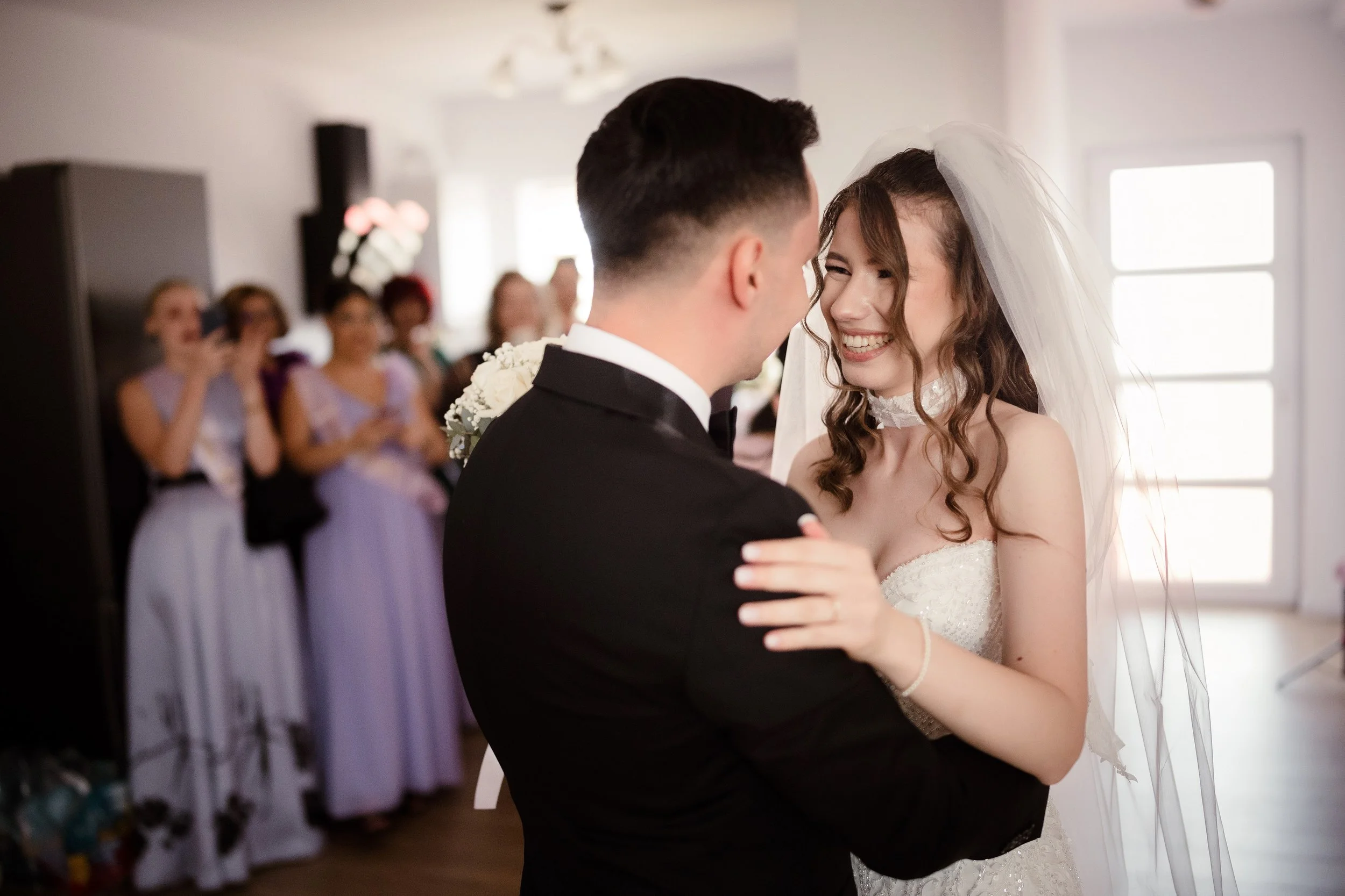 A bride and groom are dancing closely, smiling at each other in a wedding reception. The bride is wearing a white wedding dress and veil, while the groom is in a black suit with a white shirt. In the background, guests are watching and smiling, with 