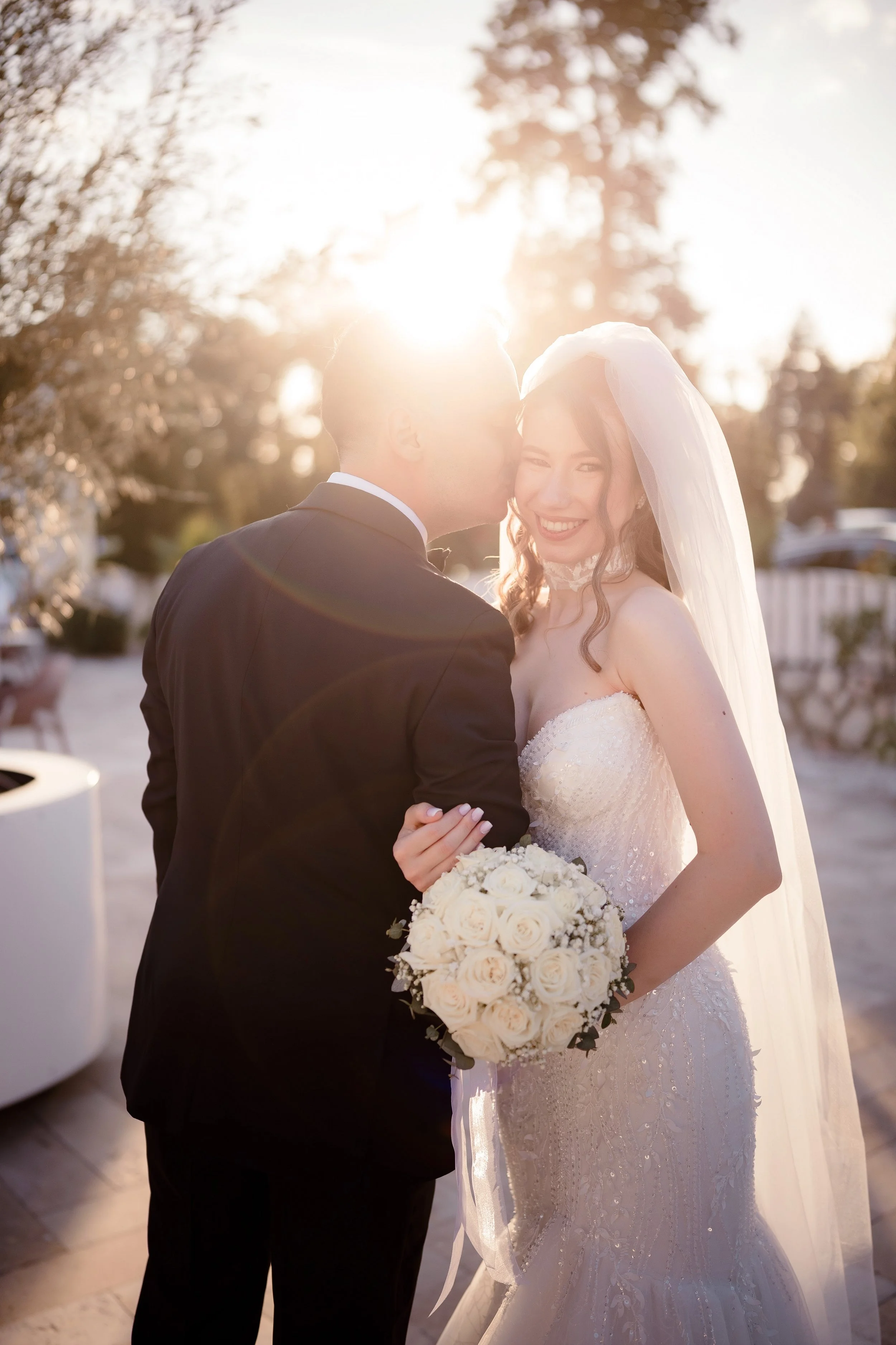 A bride and groom share a kiss outdoors during sunset. The bride is smiling and holding a bouquet of white roses, wearing a lace wedding dress and veil. The groom is dressed in a black suit. Sunlight creates a warm glow, with trees in the background.