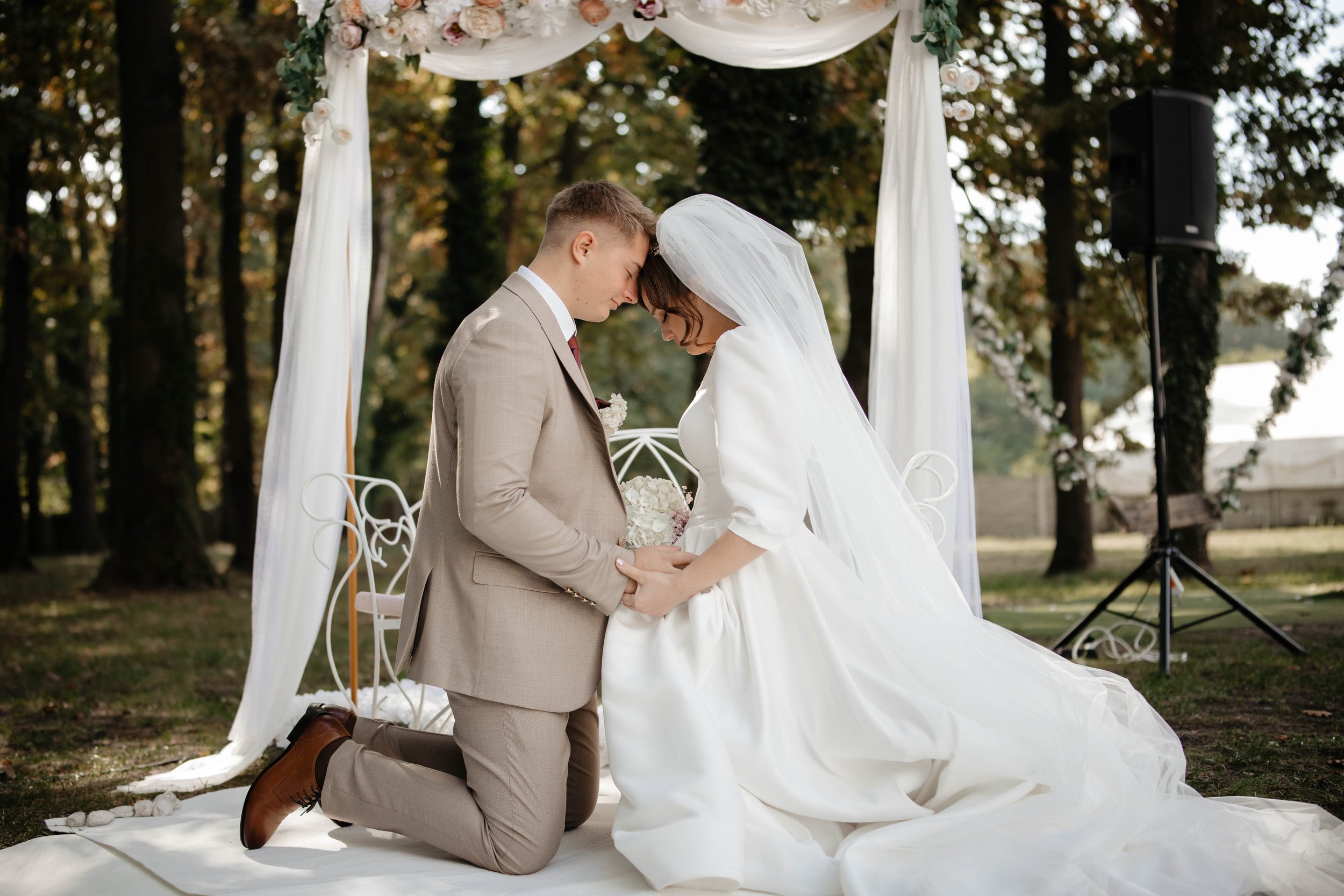 A couple getting married outdoors beneath a floral arch, with the groom kneeling and their foreheads touching, holding hands in a kneeling position, during daytime.