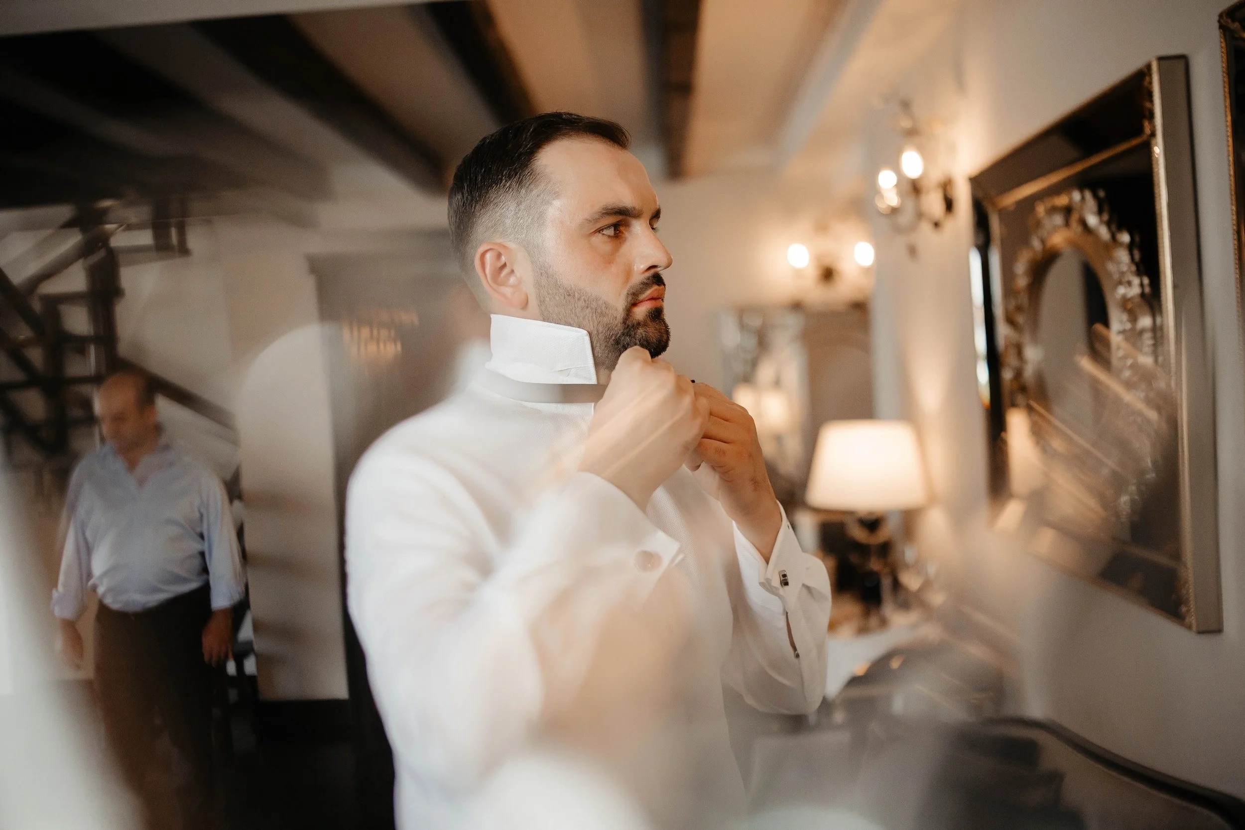 A man with a beard and styled hair adjusting his white tuxedo collar, with another man in the background, in a warmly lit room with decorative mirrors and wall sconces.