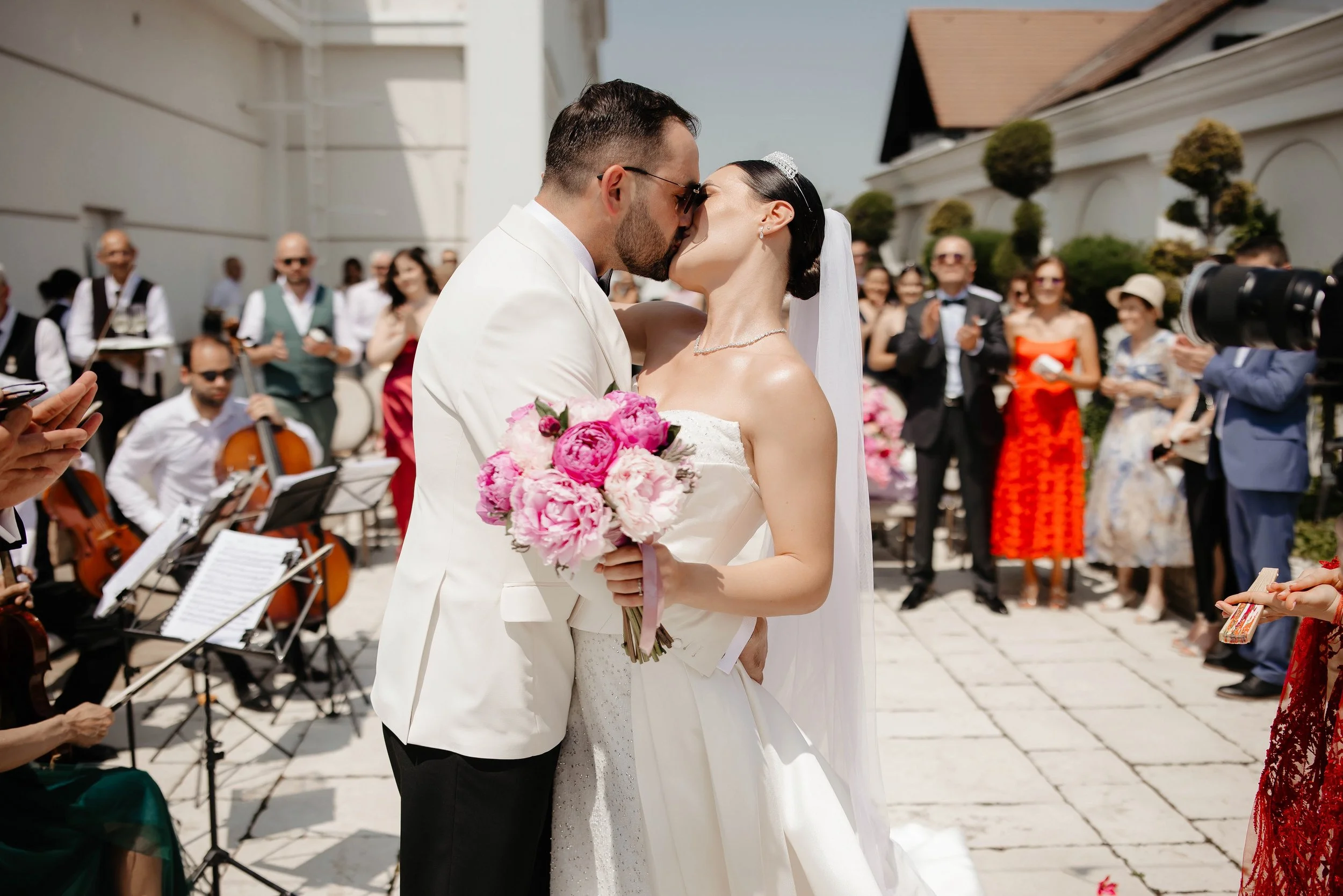 A bride and groom share a kiss during their outdoor wedding ceremony, with guests and musicians in the background.
