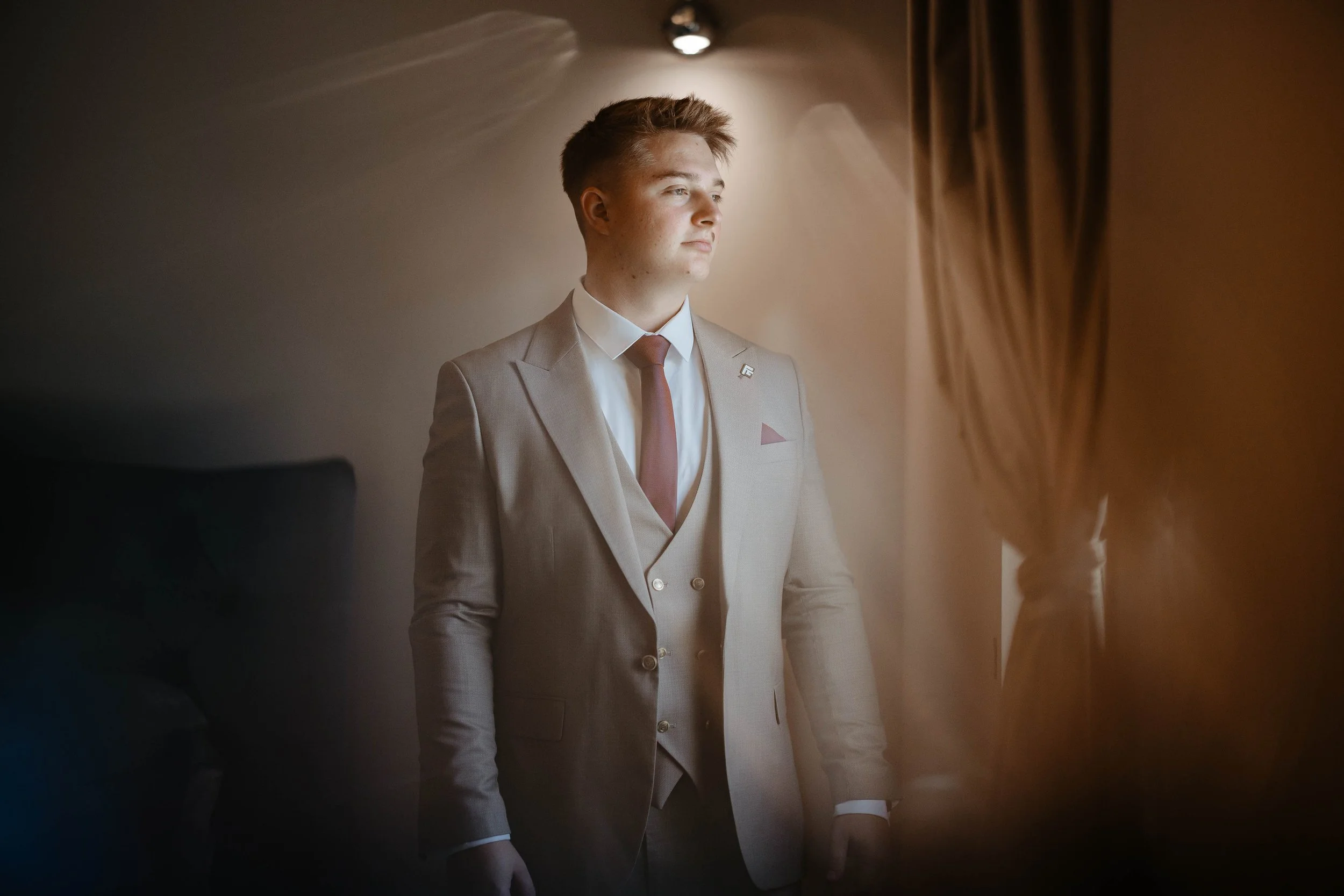 A young man in a light gray suit with a white shirt and maroon tie standing by a window, looking outside with natural light illuminating his face.