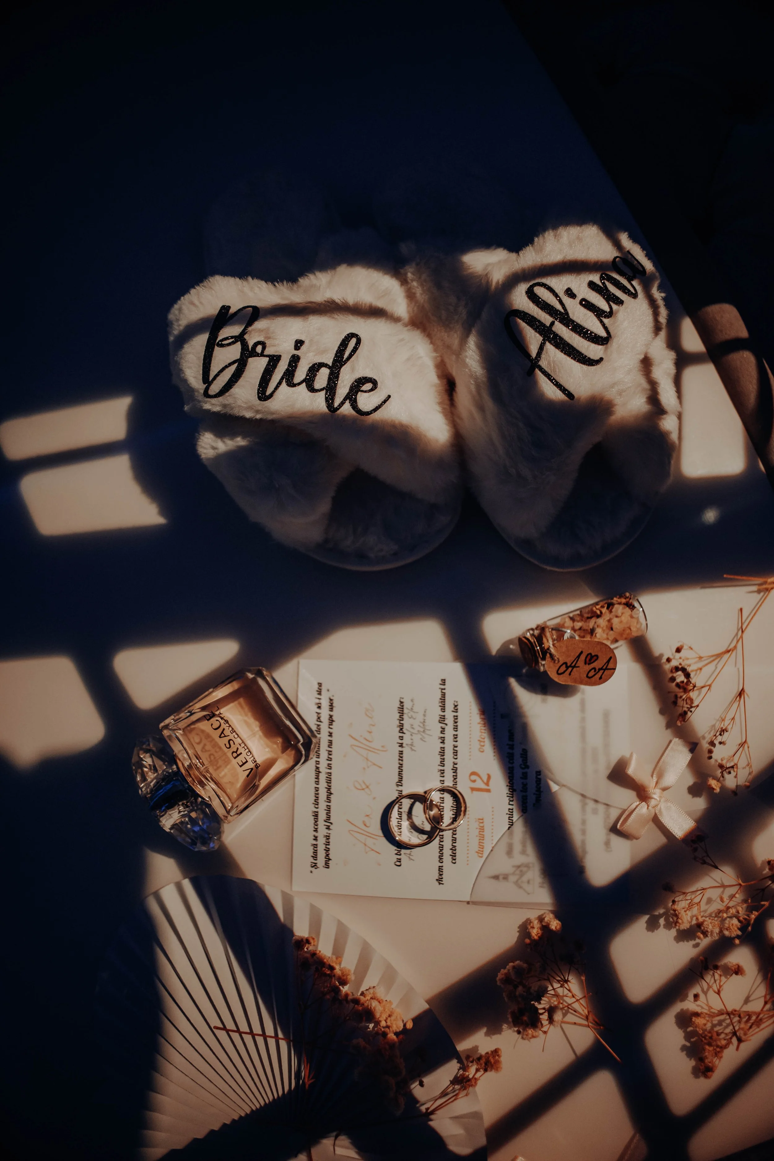 A pair of fluffy white slippers labeled 'Bride' and 'Groom' in black cursive writing, surrounded by wedding-themed items like a ring, a bottle of perfume, wedding invitations, and dried flowers, with shadows cast across the scene.