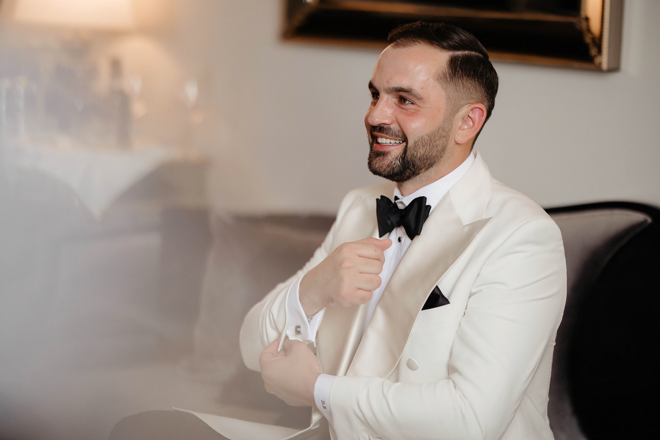A man dressed in a white tuxedo with a black bow tie, smiling and adjusting his tuxedo at a formal event, seated indoors.