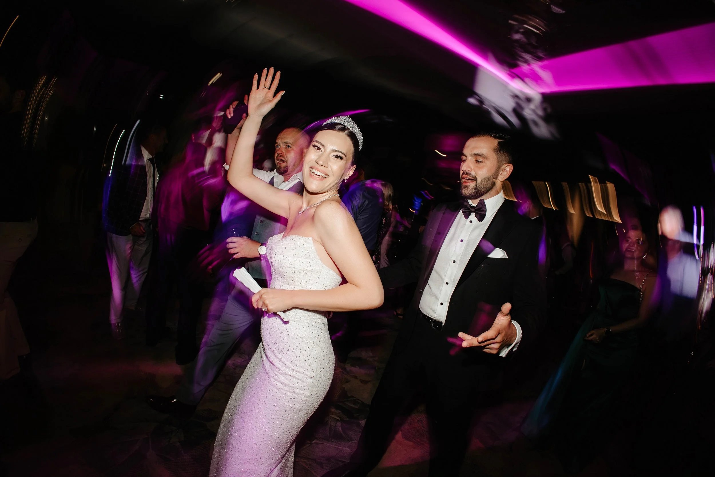 A woman in a white strapless wedding gown and tiara dancing and smiling at a wedding reception, with a man in a tuxedo next to her, and other guests dancing in the background.